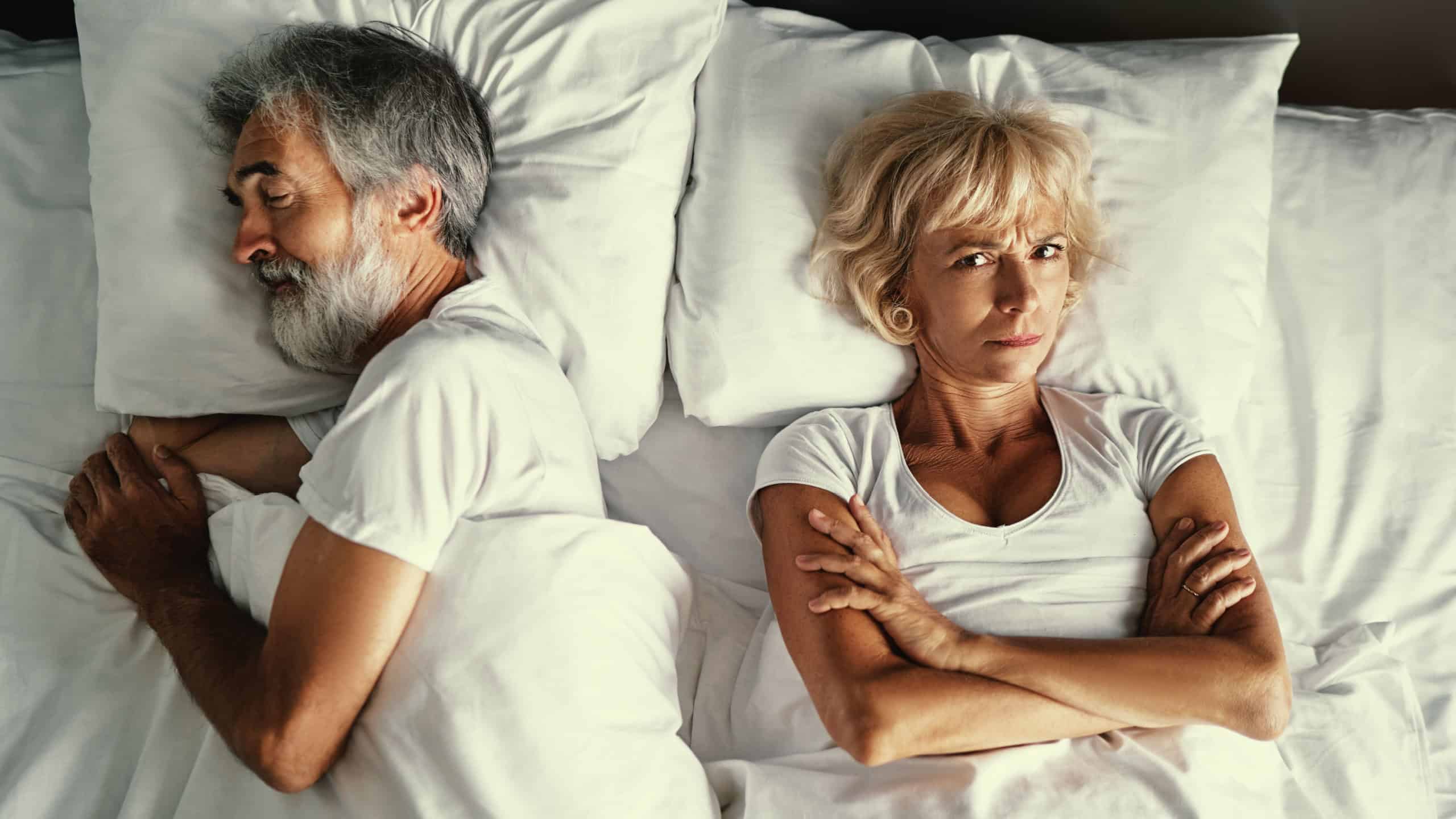 Overhead of senior couple in the bed. Pretty aged woman is angry with her sleeping husband.