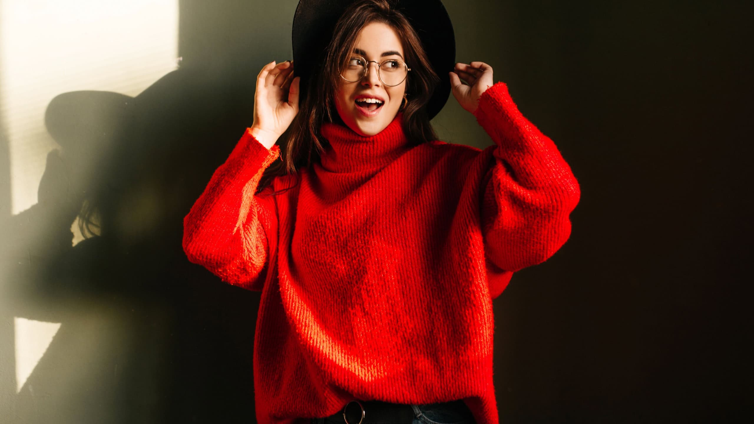 Fashion-monger in wide-brimmed hat and glasses is posing in studio against background of her own shadow. Girl in red oversize sweater looks aside