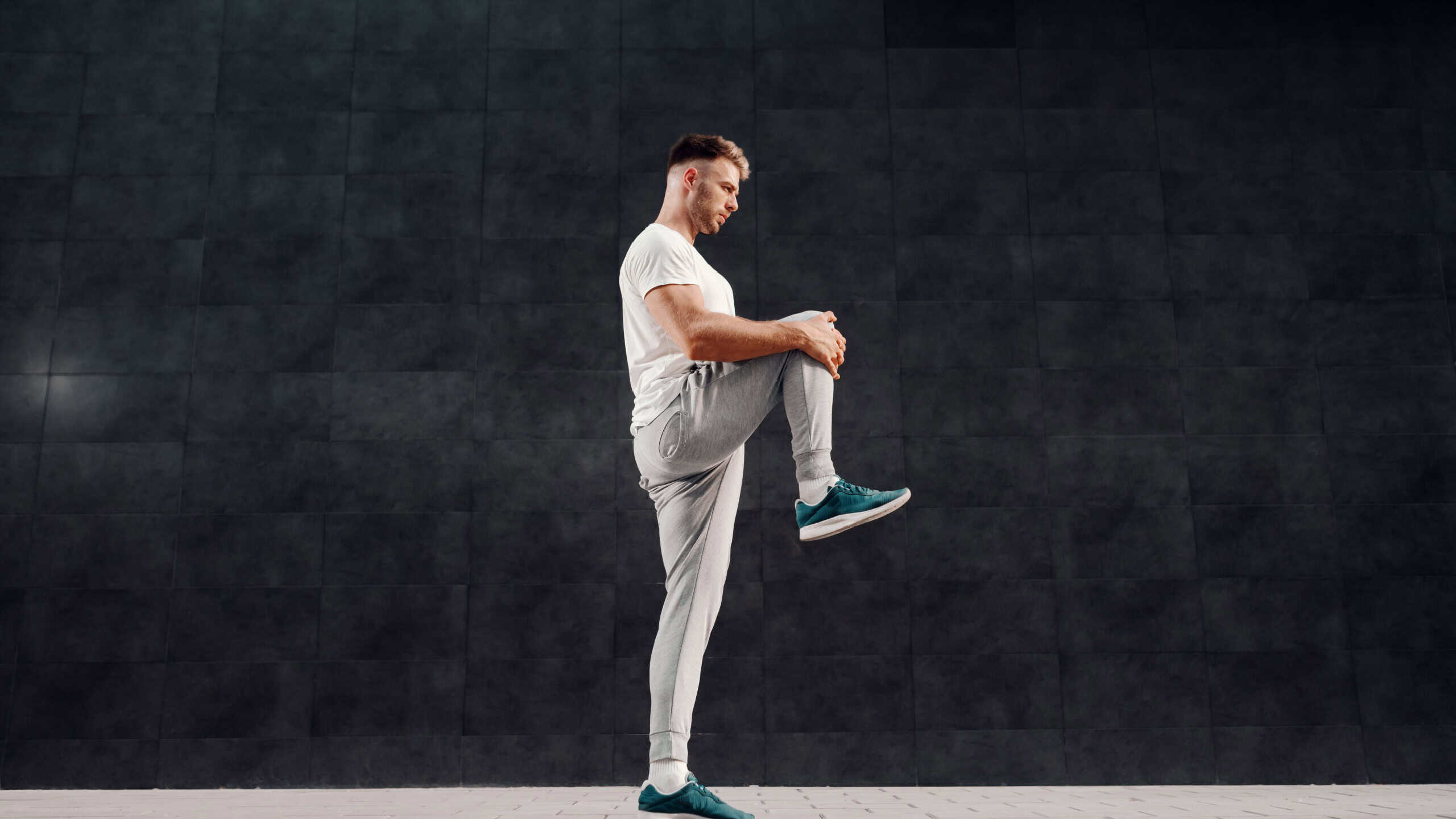 Full length of attractive Caucasian bearded blonde man standing on one foot and stretching leg. In background is gray wall.