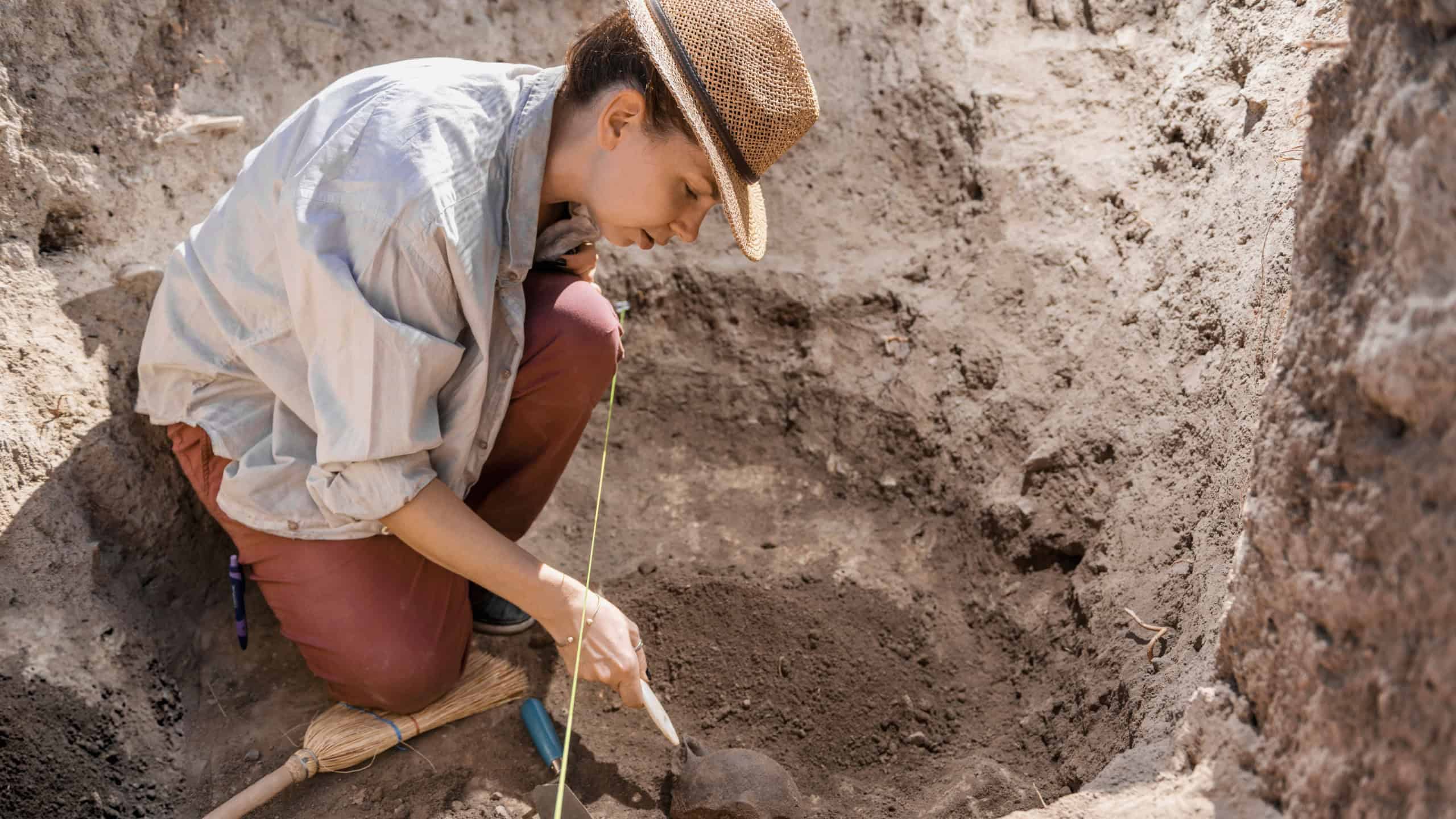 Archaeologist digging with hand trowel, recovering ancient pottery object from an archaeological site.
