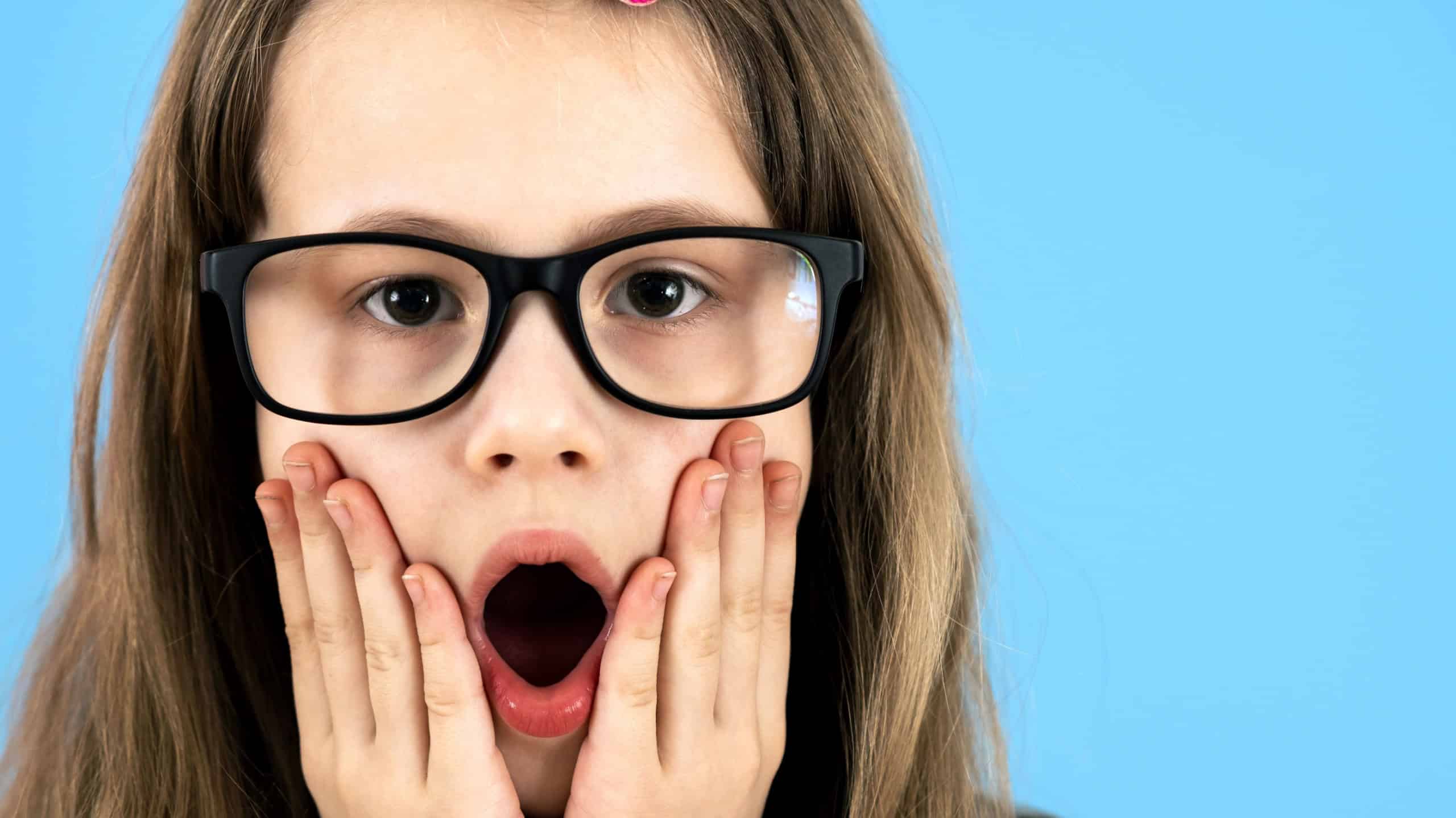 Portrait of surprised child girl wearing looking glasses holding hands to her face isolated on blue background.