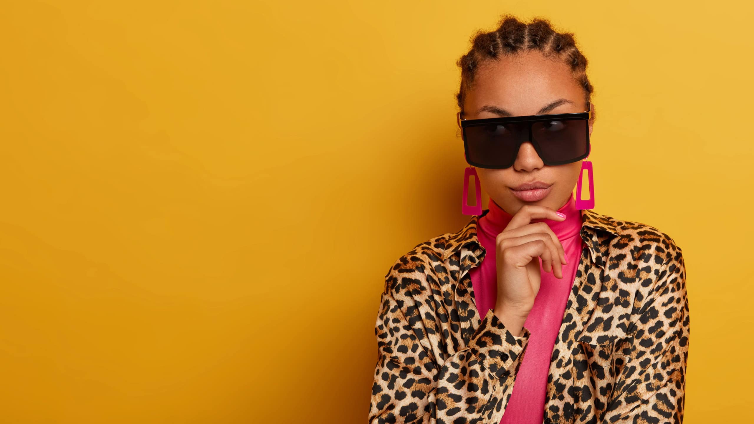 Spring fashion concept. Elegant young woman dressed in latest style outfit, stands thoughtfully, holds hand on chin, concentrated aside, looks through big sunglasses, isolated on yellow background
