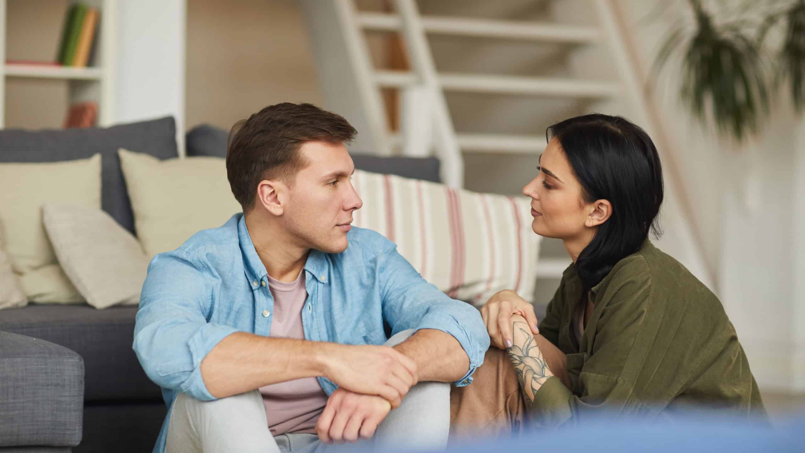 Warm toned portrait of modern young couple talking to each other sincerely while sitting on floor in cozy home interior, copy space