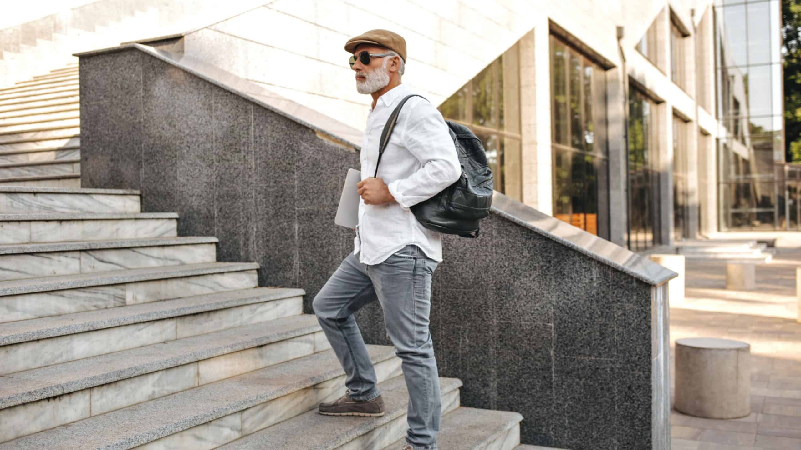 Portrait of man in grey jeans and white shirt walking in city. Modern bearded guy in stylish clothes climbs stairs and holds black backpack..