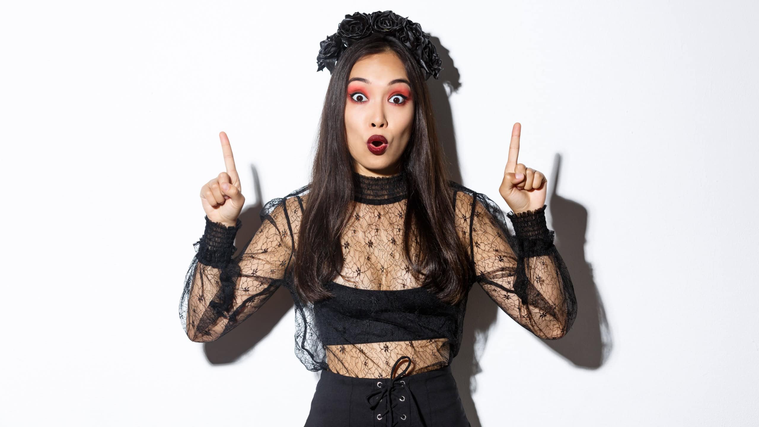 Surprised asian woman in halloween costume showing amazing promo offer for halloween, pointing fingers up and looking amazed, standing in black gothic dress with mysterious makeup