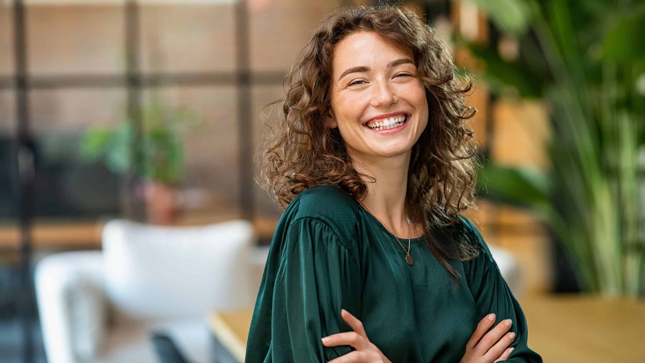 Portrait of young smiling woman looking at camera with crossed arms. Happy girl standing in creative office. Successful businesswoman standing in office with copy space.