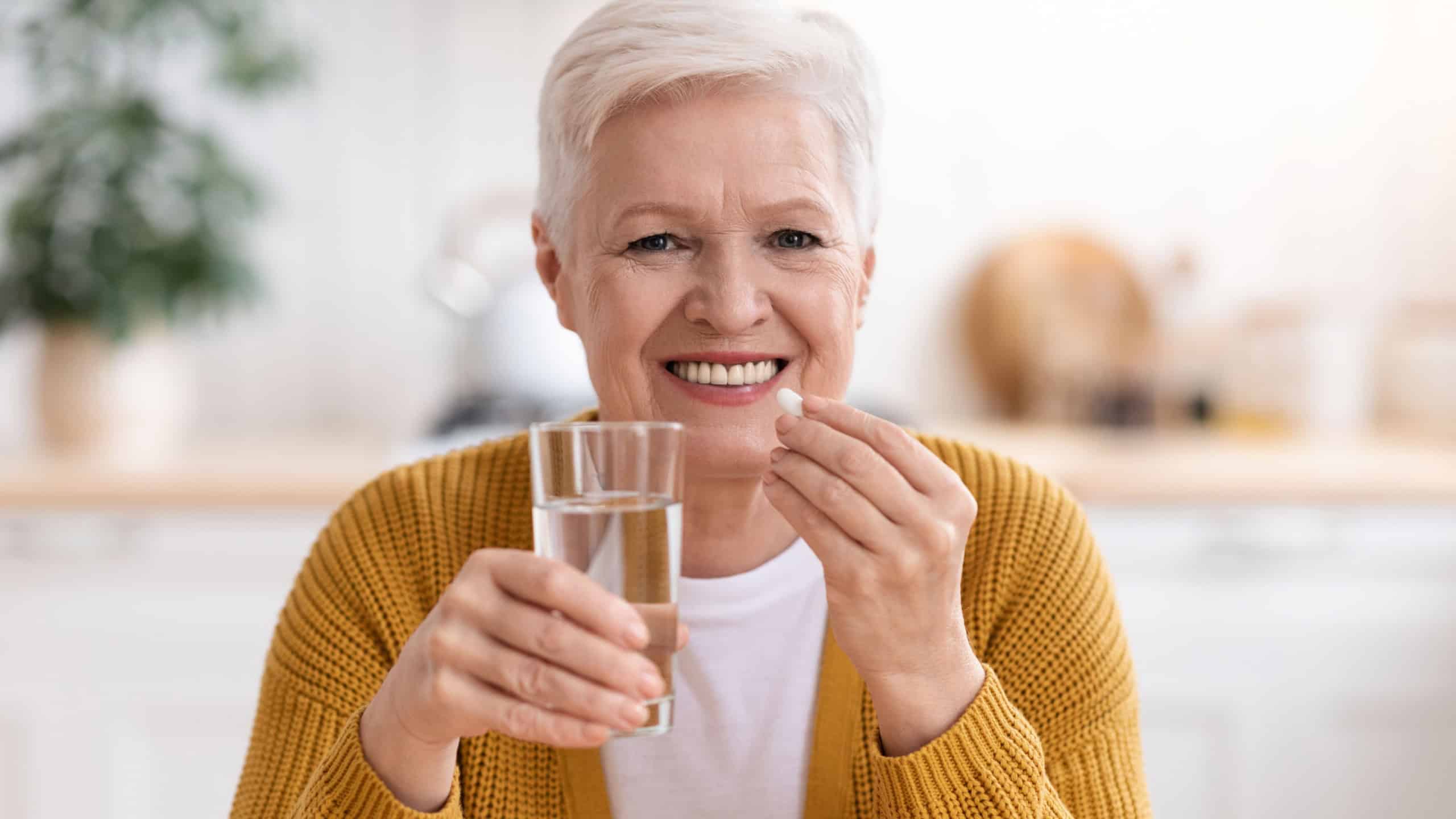 Supplements, vitamins, healthy lifestyle concept. Closeup of happy senior lady holding glass of water and taking pill, kitchen interior, copy space. Smiling grey-haired elderly woman using supplements