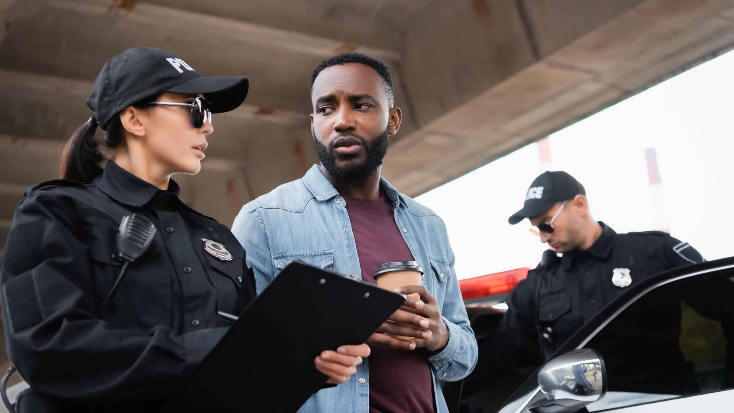 Policewoman with clipboard talking with african american victim with takeaway coffee near colleague and car on blurred background