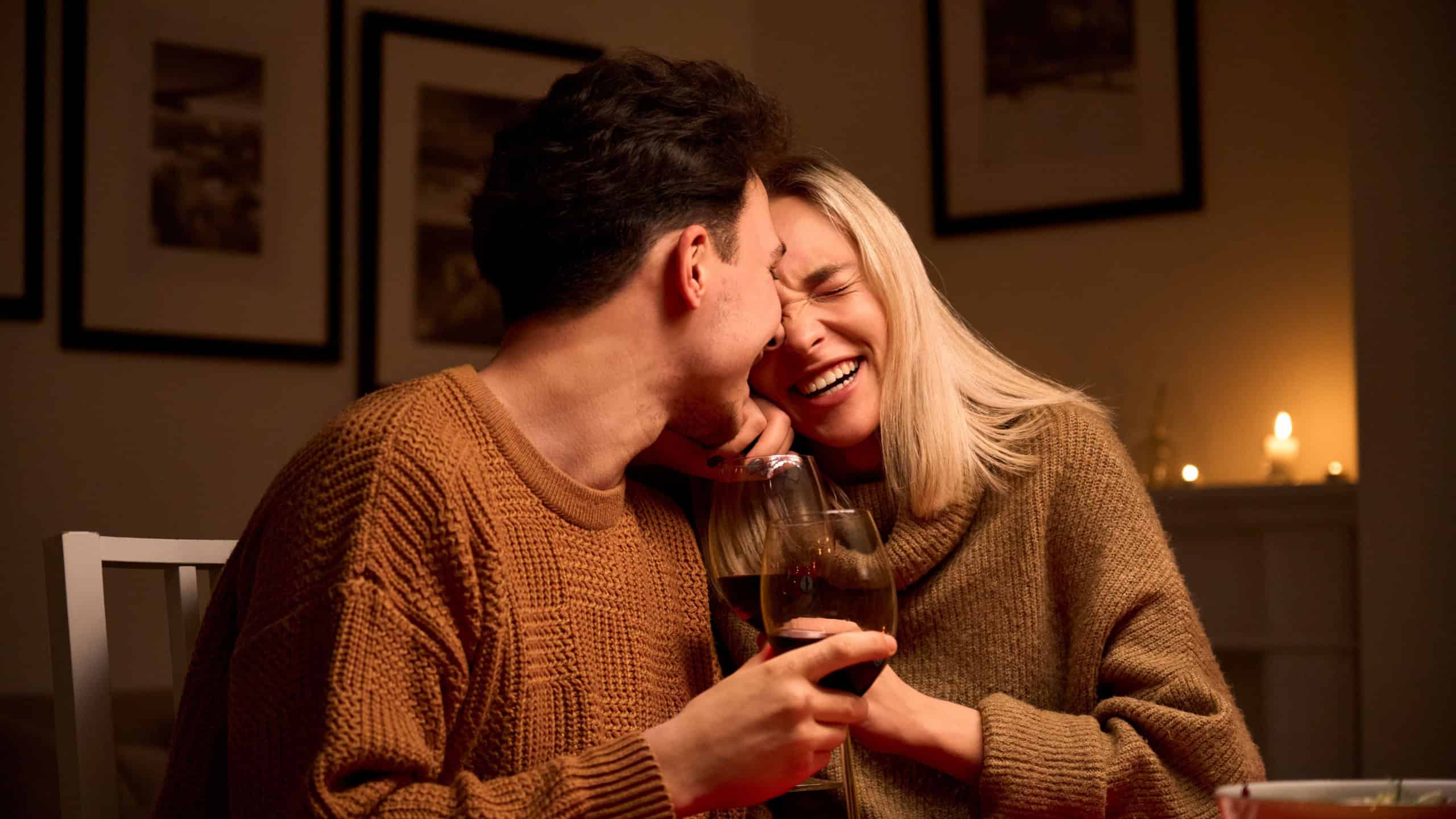 Happy young couple in love hugging, laughing, drinking wine, enjoying talking, having fun together celebrating Valentines day dining at home, having romantic dinner date with candles sitting at table.