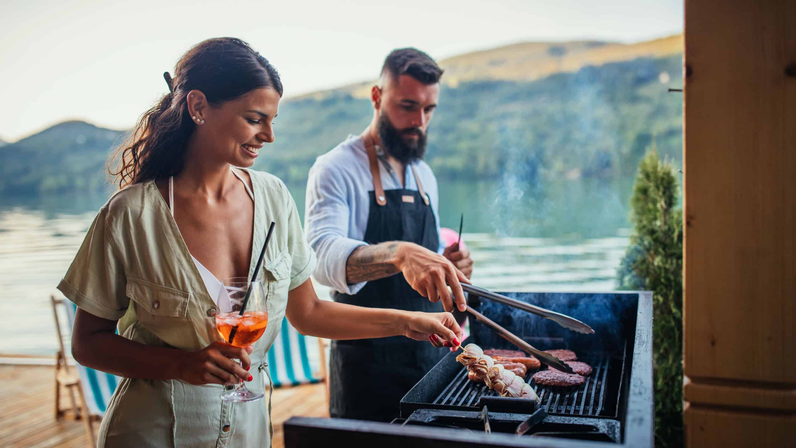 Happy young couple barbecuing meat on the grill while standing at the terrace of weekend cottage near lake