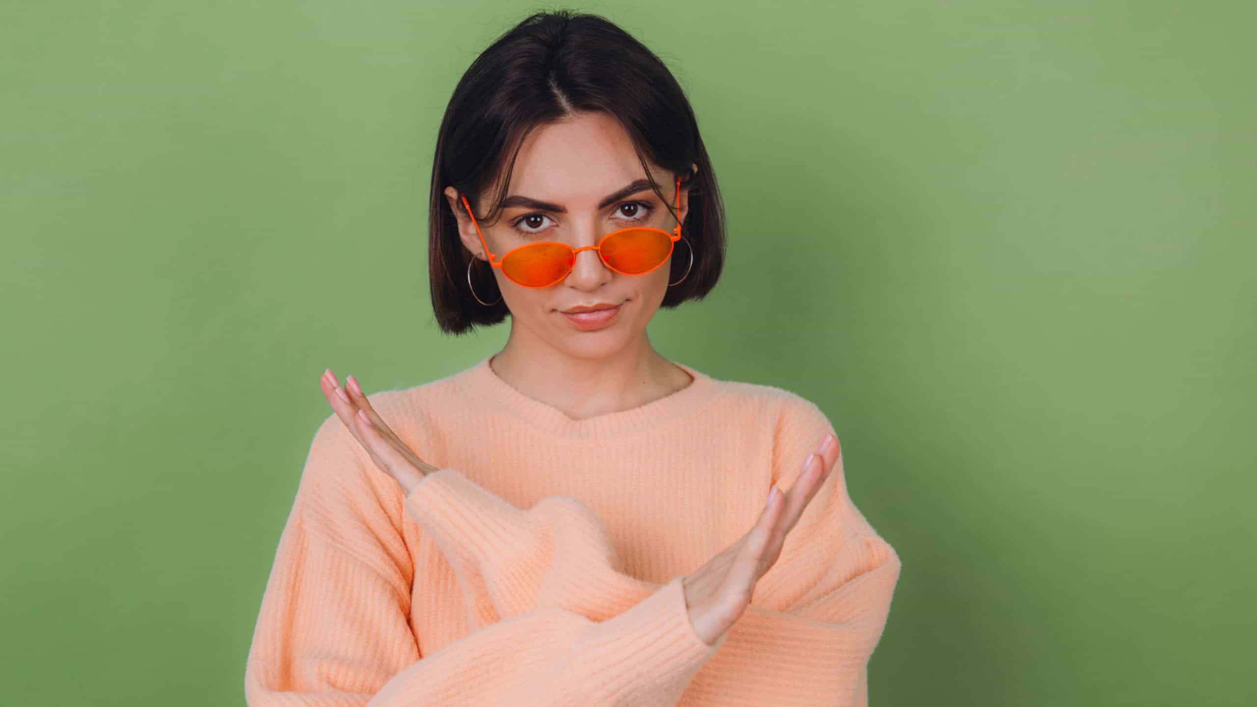 Young woman in casual peach and orange eyeglasses sweater isolated on green olive background serious showing stop gesture with crossed hands copy space