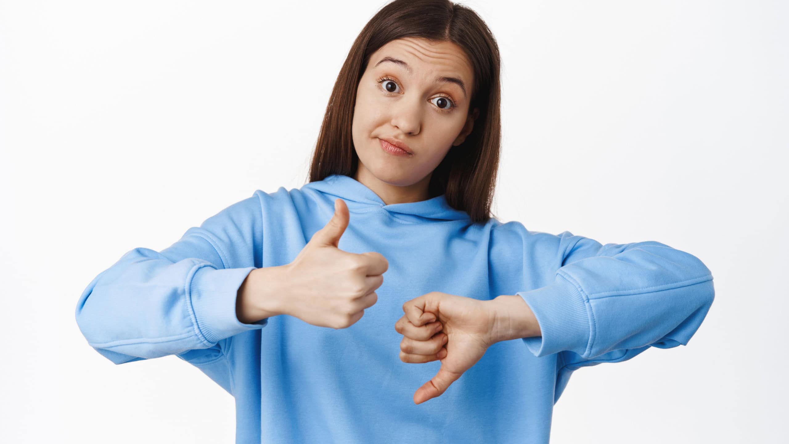 Not bad. Young woman smirk, shows like dislike, thumbs up and thumbs down gesture, medium rating, average result, standing in hoodie against white background