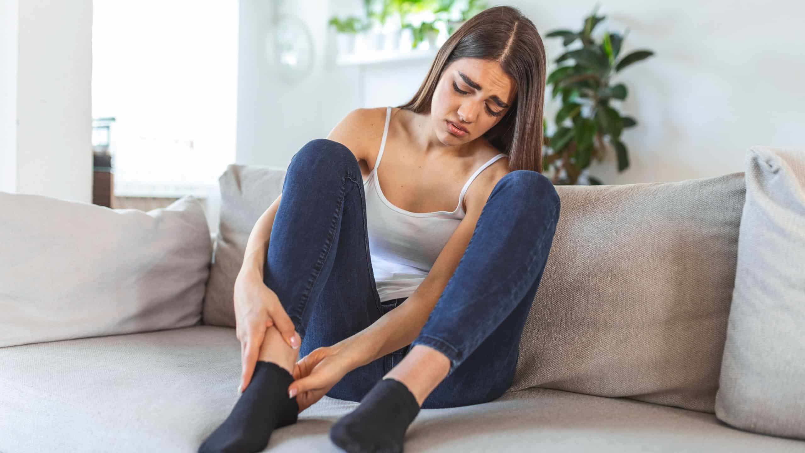 Hands massaging swollen foot while sitting on sofa during the day at home. Photo of Young Caucasian woman suffering from pain in leg. Woman massaging her legs after all day at work in office