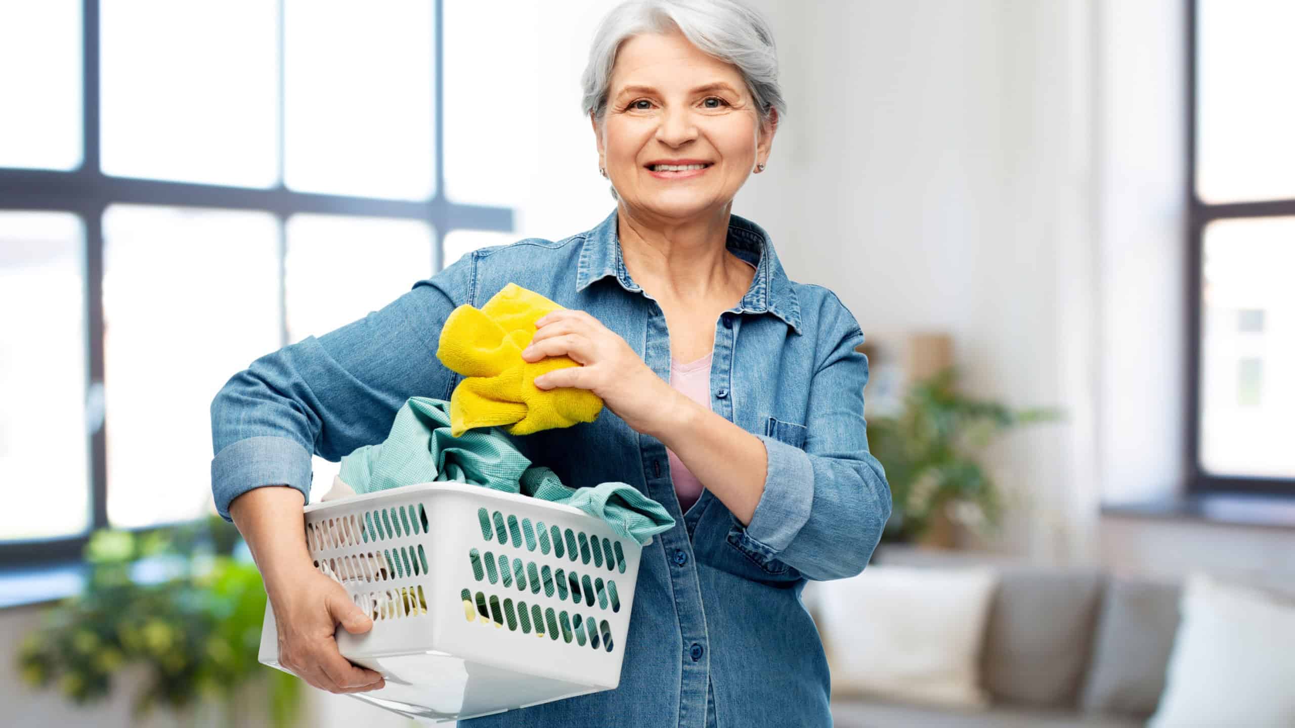 cleaning, wash and old people concept - portrait of smiling senior woman in denim shirt with towels in laundry basket over home background