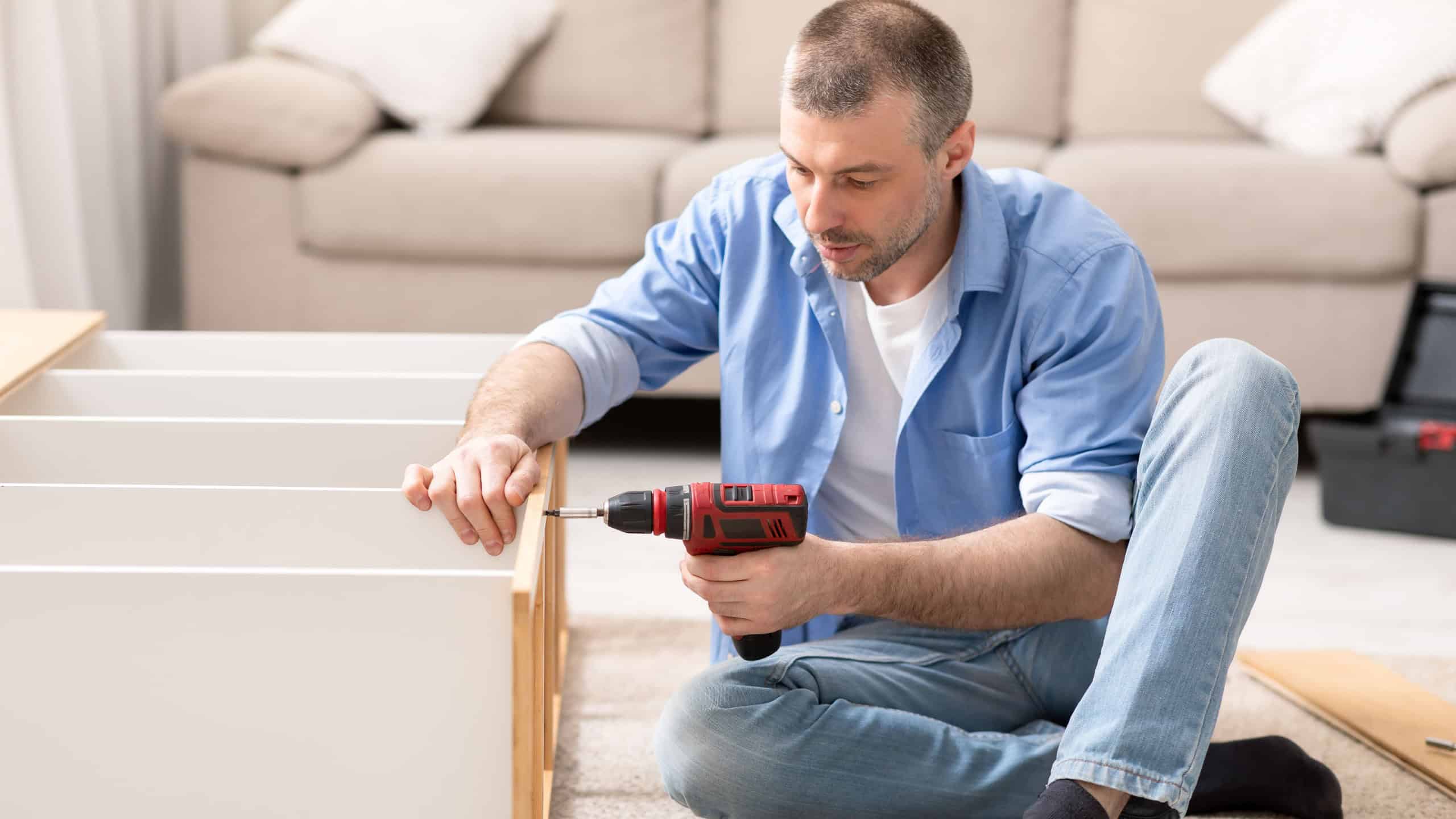 Mature Man Assembling Shelf Using Electric Drill Tool Sitting On Floor At Home. Male Housework And House Furnishing. Handyman Fixing Wooden Cabinet During Renovation