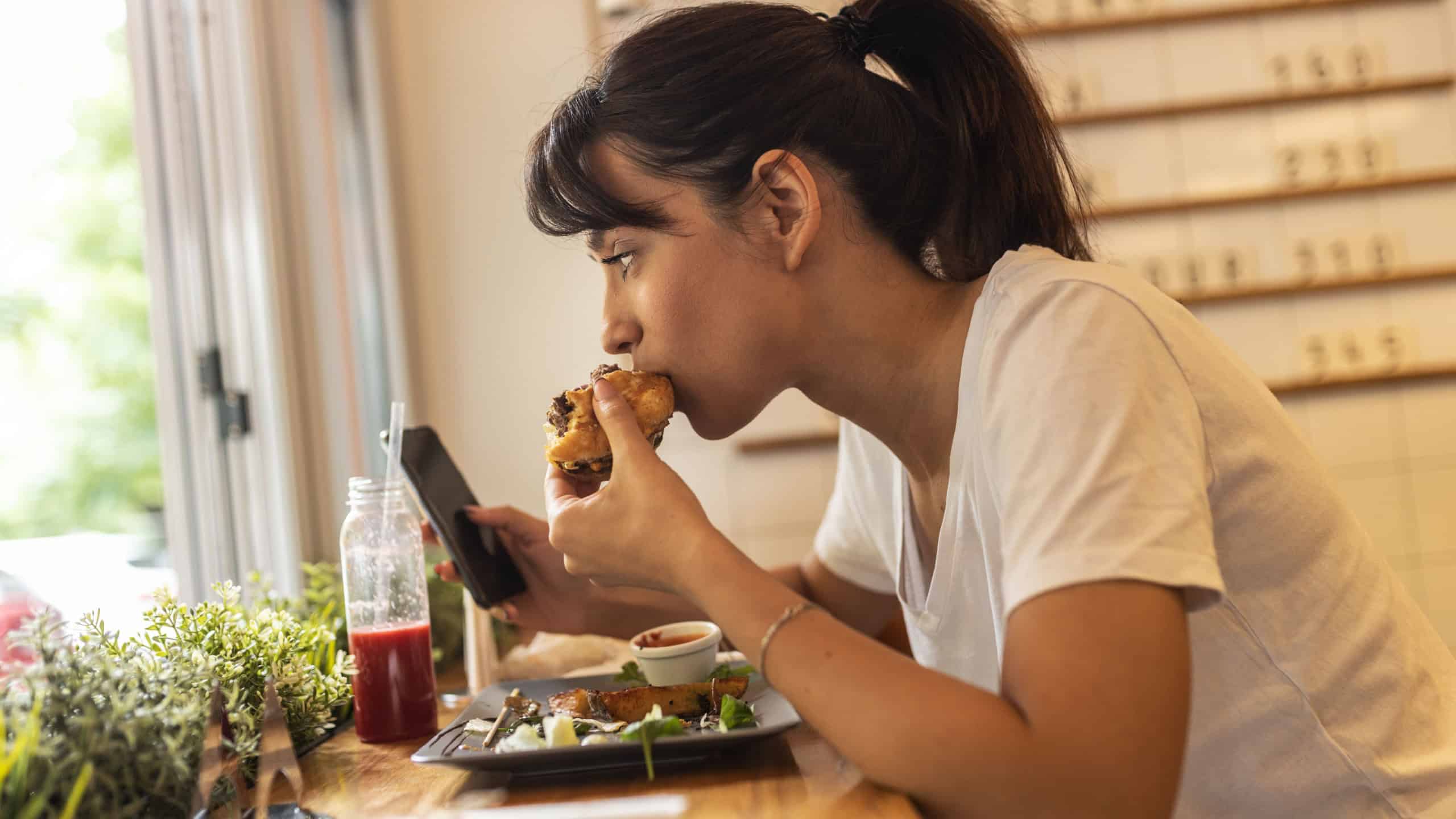 Teenage girl sitting in fast food restaurant eating burger and drinks fresh organic juice.