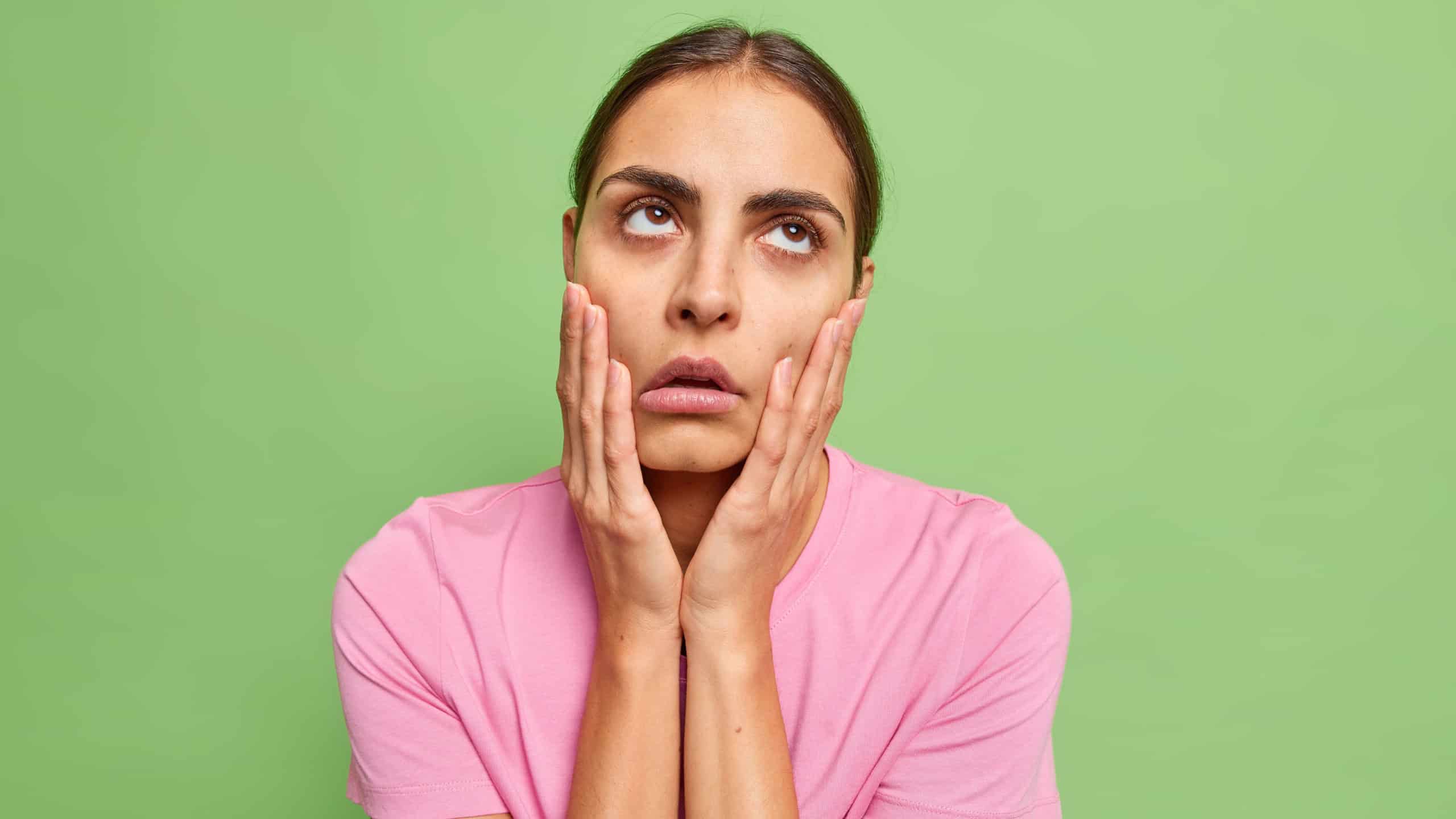 Uninterested bored young woman keeps hands on cheeks fed up of dull conversation looks above makes facepalm and rolls eyes hears nonsense wears casual t shirt isolated over green background.