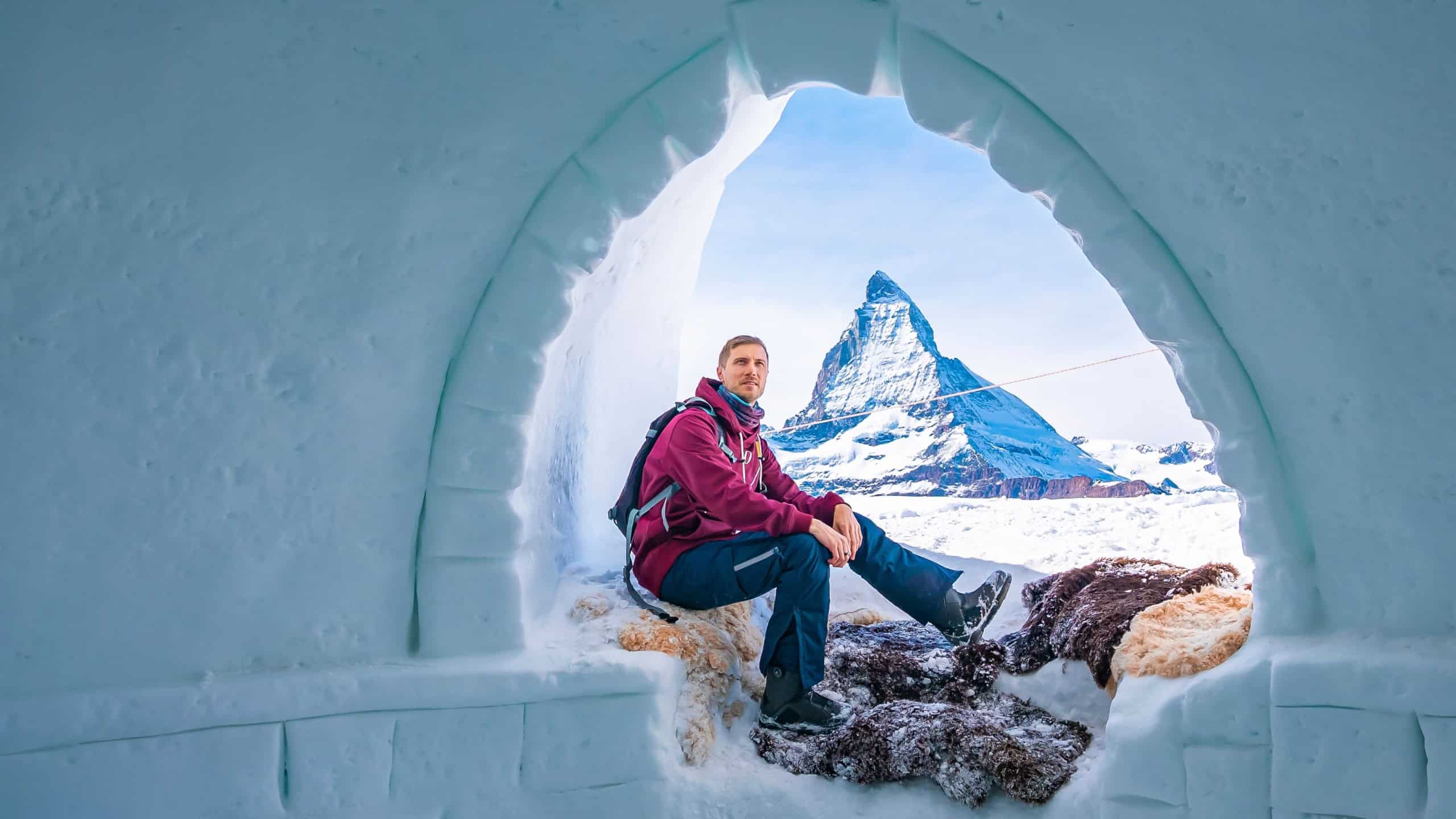 Portrait of young tourist resting at entrance of igloo. Famous snowcapped matterhorn peak is in background. Man enjoying winter in alpine region.