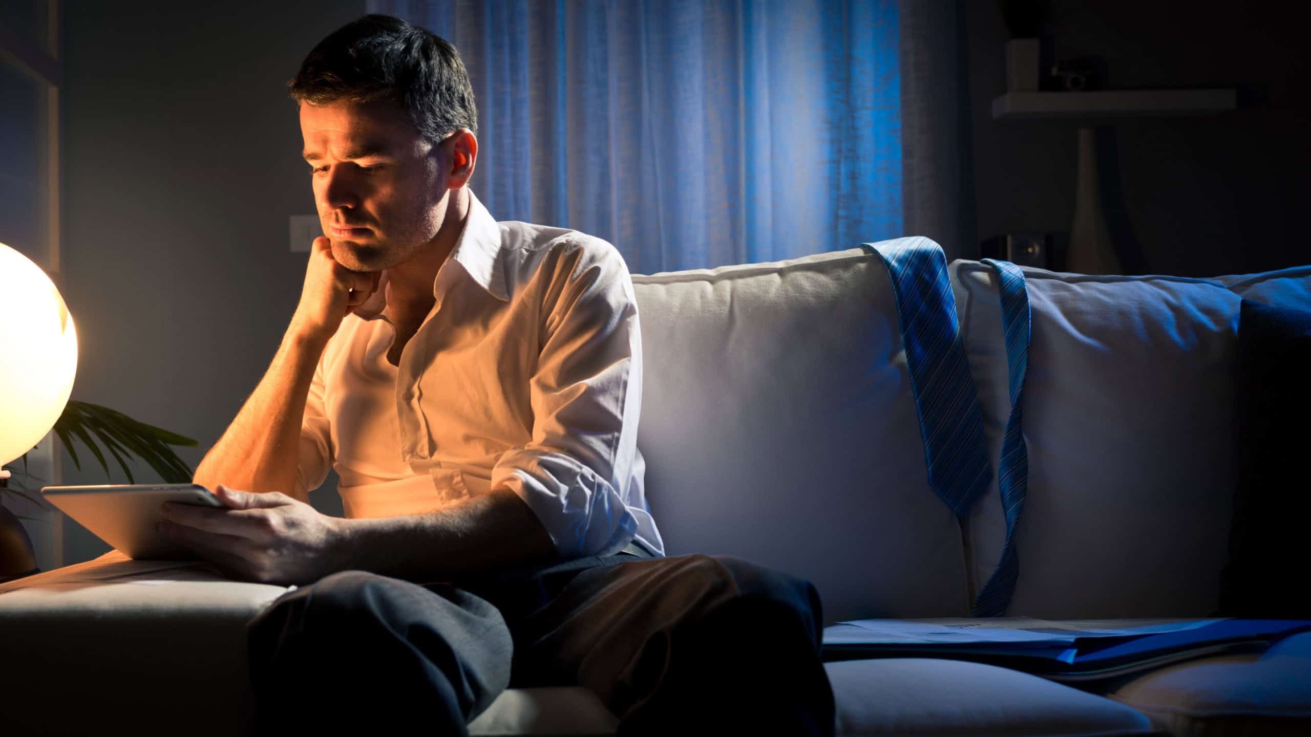 Businessman working overtime late at night in the living room, with tablet and paperwork.