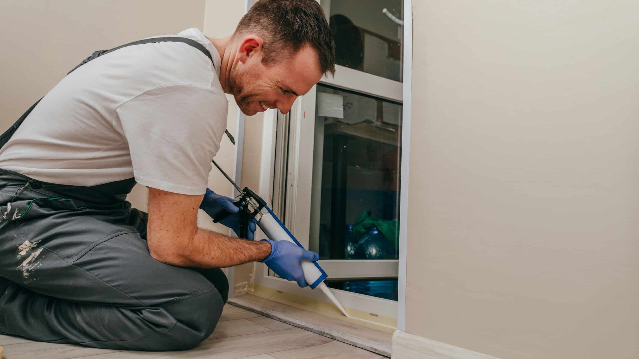 Young man wearing overalls sealing cracks between door and trim using waterproof silicone caulk on the balcony.