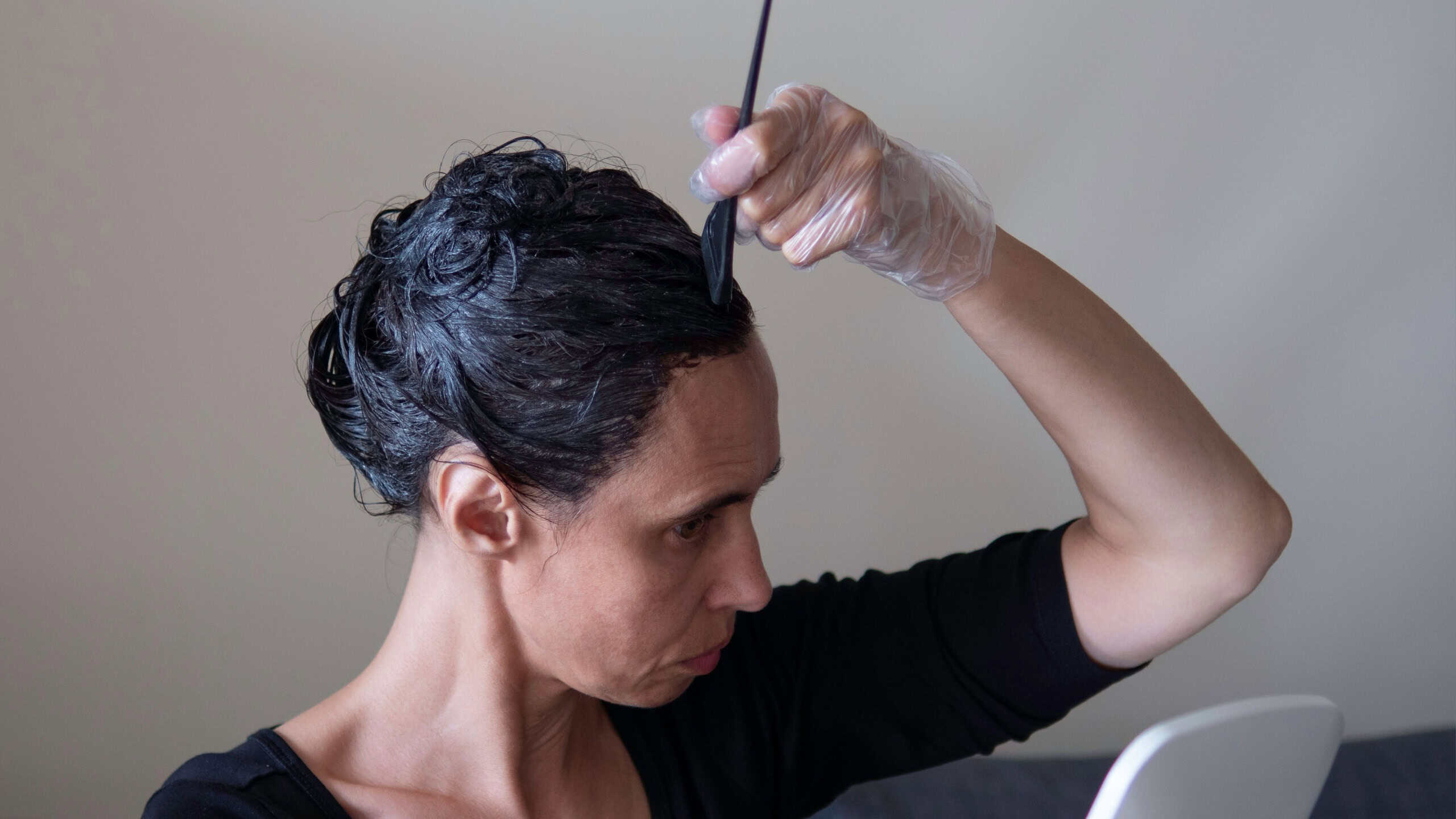 Middle age woman colouring dark hair with gray roots at home. Woman sitting on couch and dyeing hair using black brush and looking to the mirror