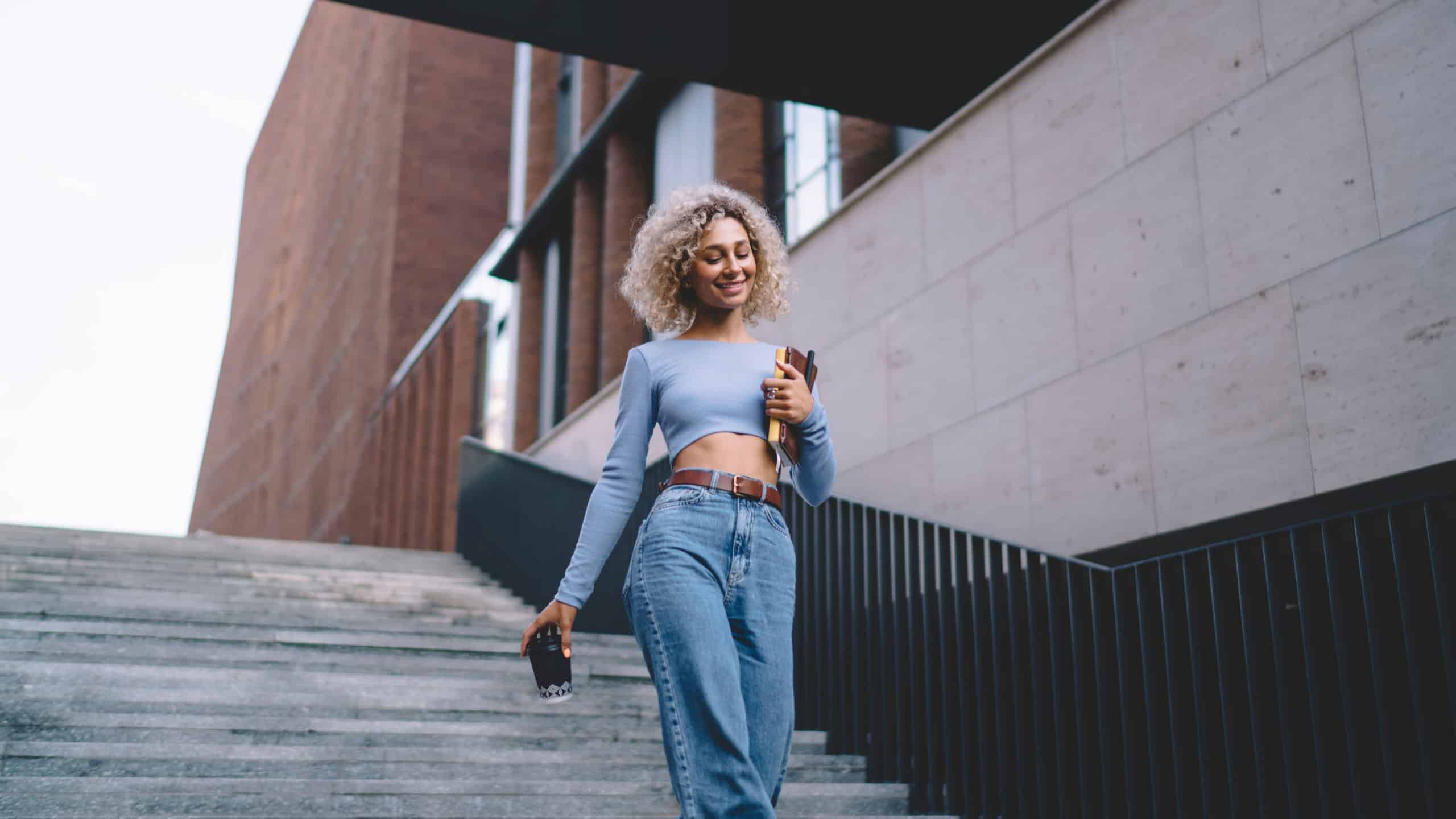 Low angle of cheerful positive young female student in casual clothes smiling and looking away while walking down steps near railing