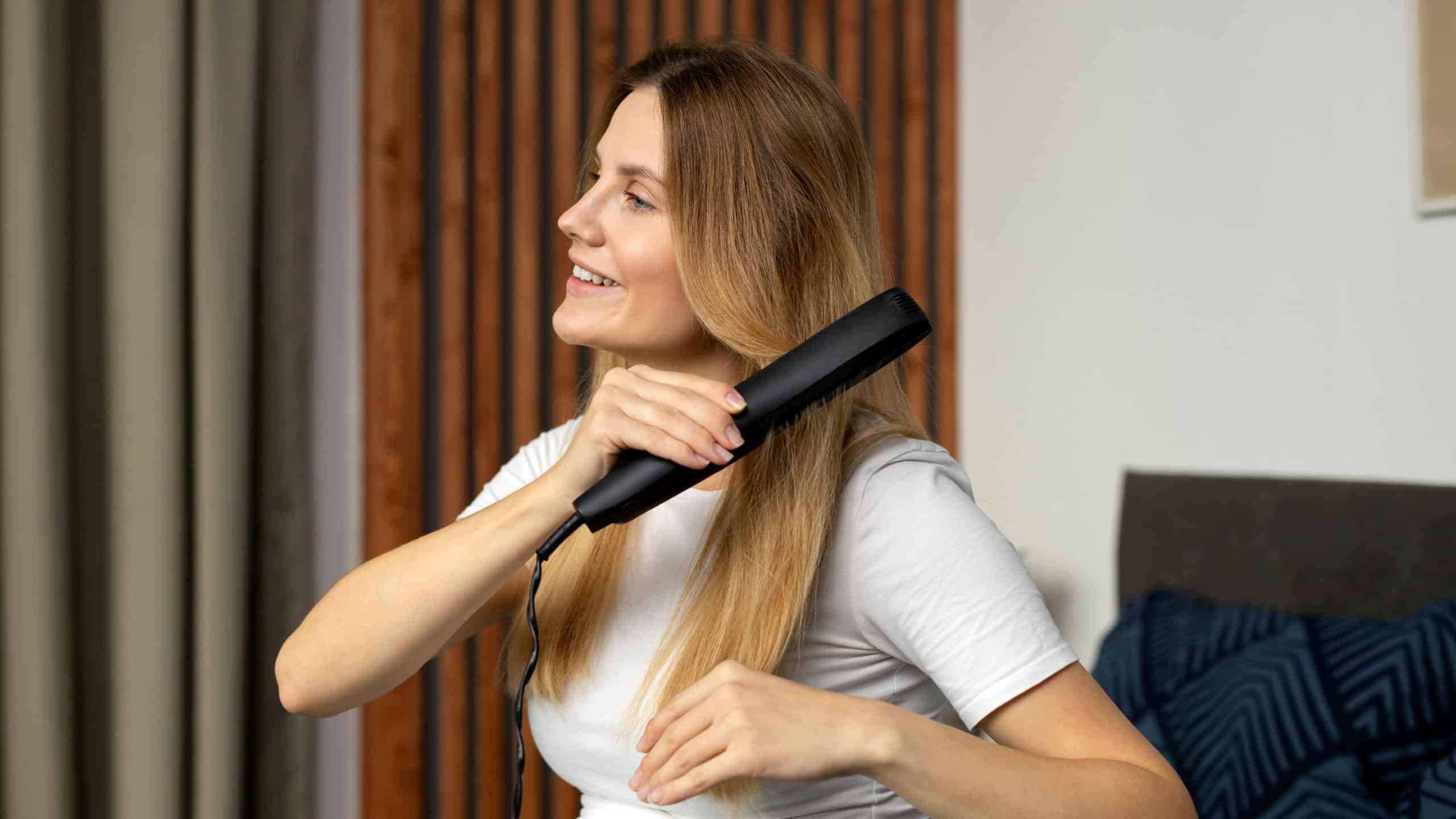 Pregnant beautiful woman in white t shirt is using hair straightener and smiling while relaxing at home. Beauty routine concept 