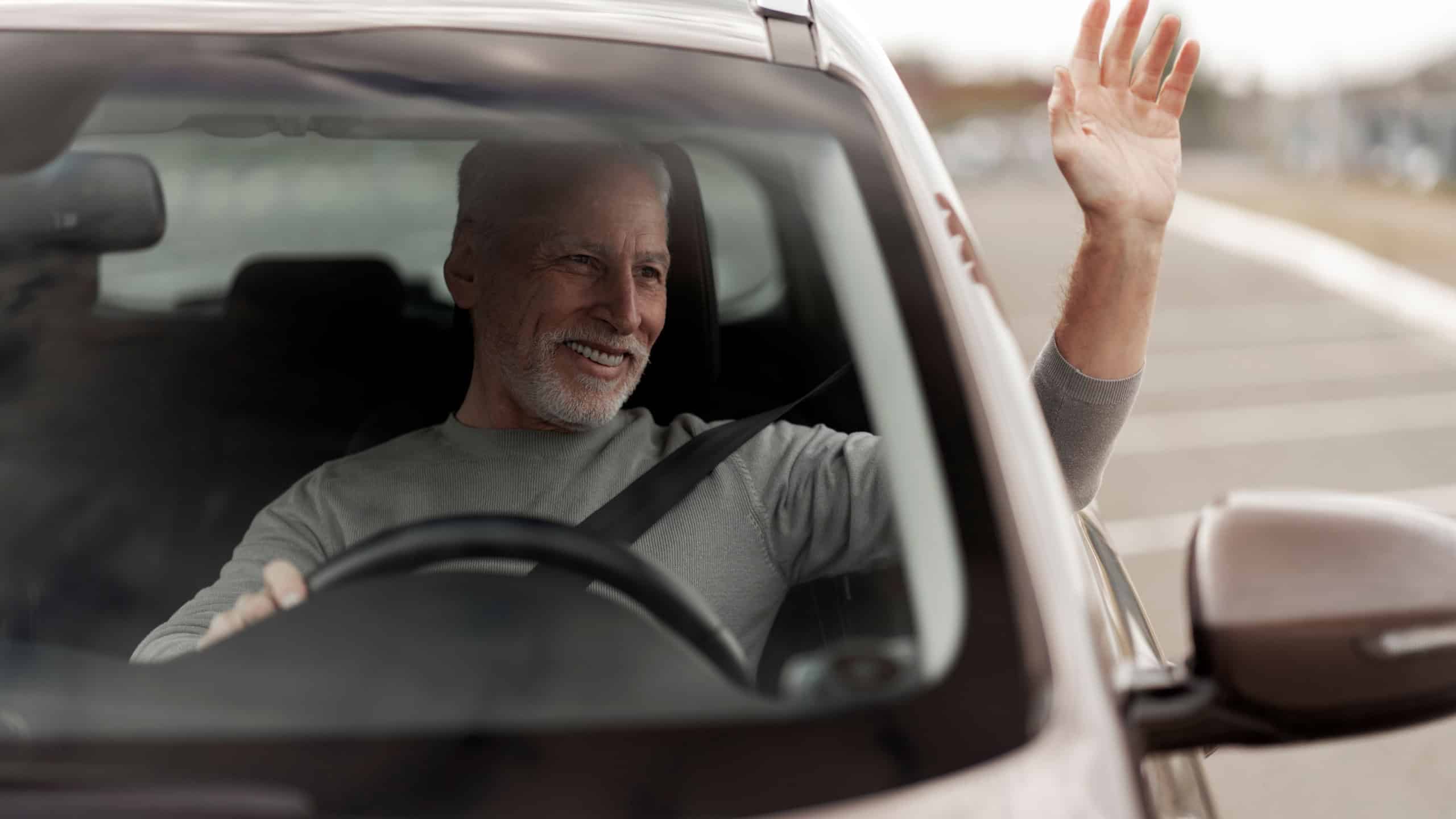 View through the windshield to a handsome senior man, driver, smiling a cheerful toothy smile, greeting, waving with hand while driving his new automobile. Lifestyle. Success. Buying new car
