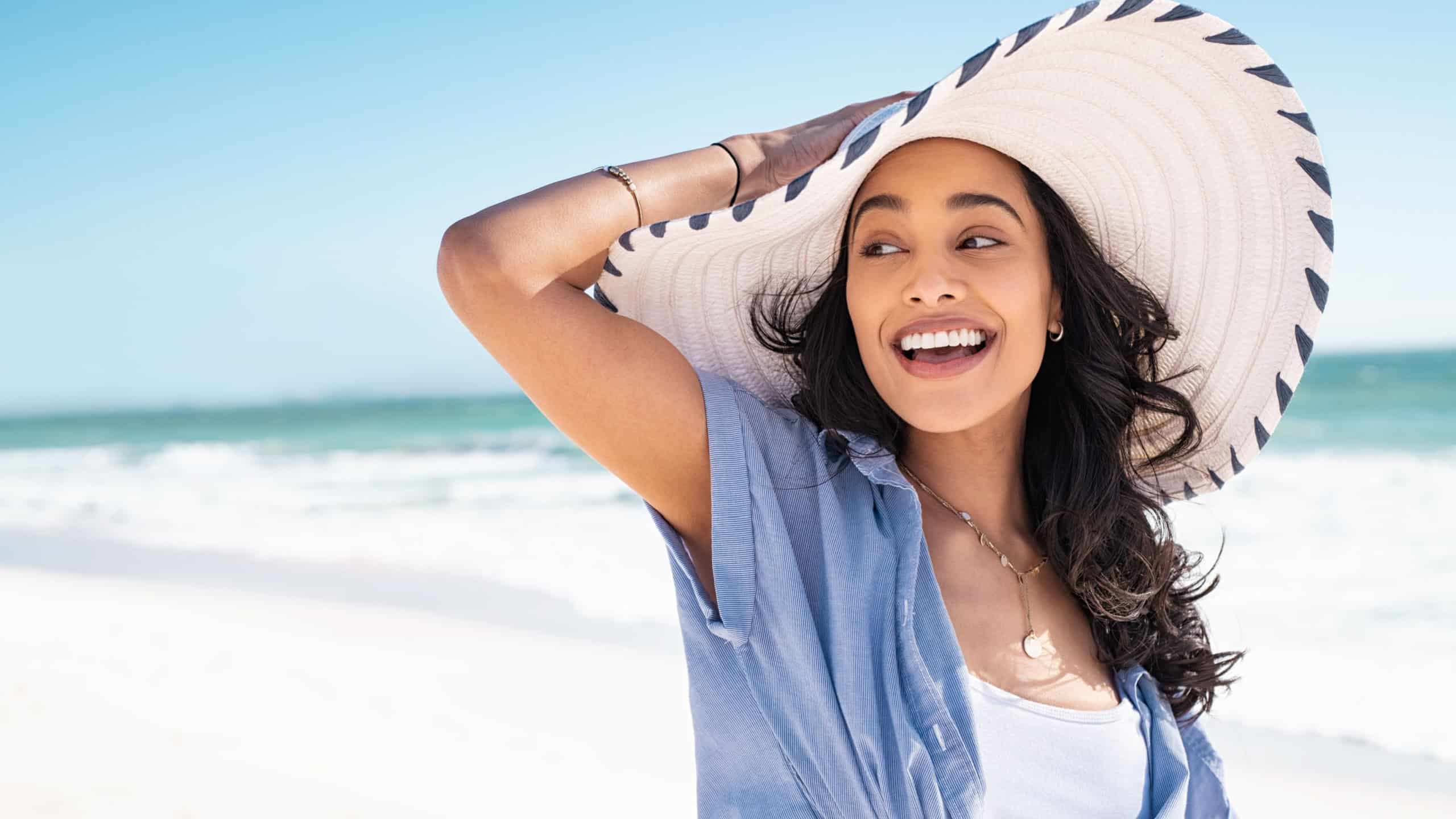 Carefree woman with white and blue straw hat walking at beach. Young smiling latin woman on vacation enjoying sea breeze. Attractive hispanic girl relaxing at beach with the sea behind.