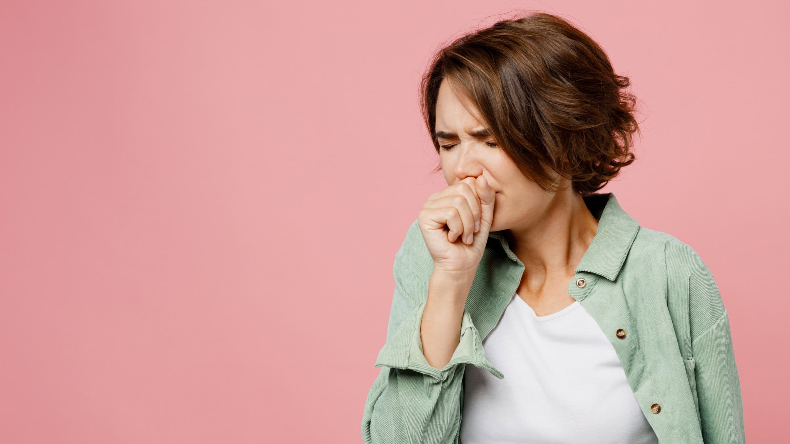 Young sick ill woman 20s she wearing green shirt white t-shirt cough hold hand near mouth isolated on plain pastel light pink background studio portrait. People healthcare pandemic lifestyle concept