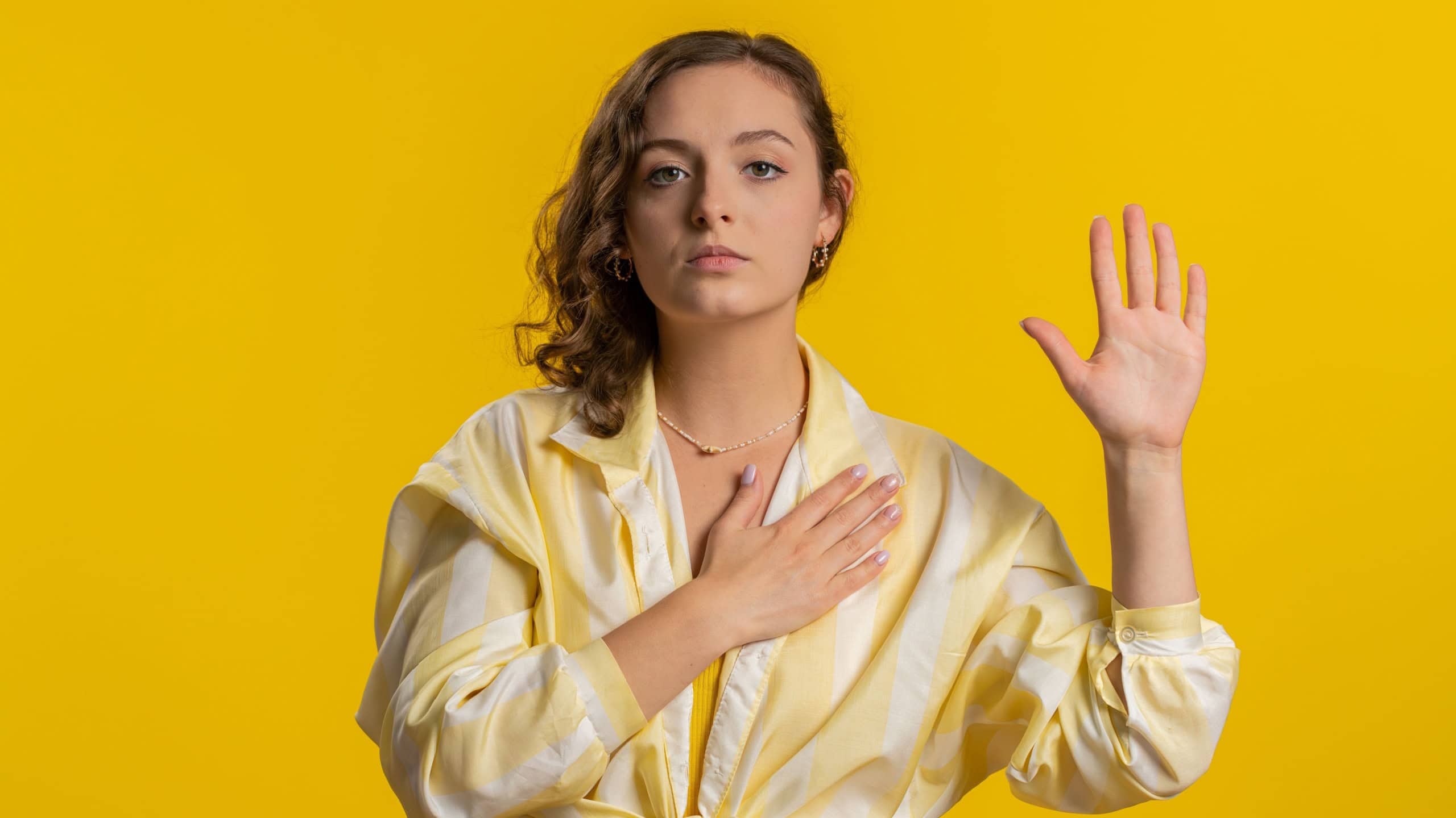 I swear to be honest. Sincere responsible young woman raising hand to take oath, promising to be honest and to tell truth, keeping hand on chest. Girl isolated on yellow studio background. Lifestyles