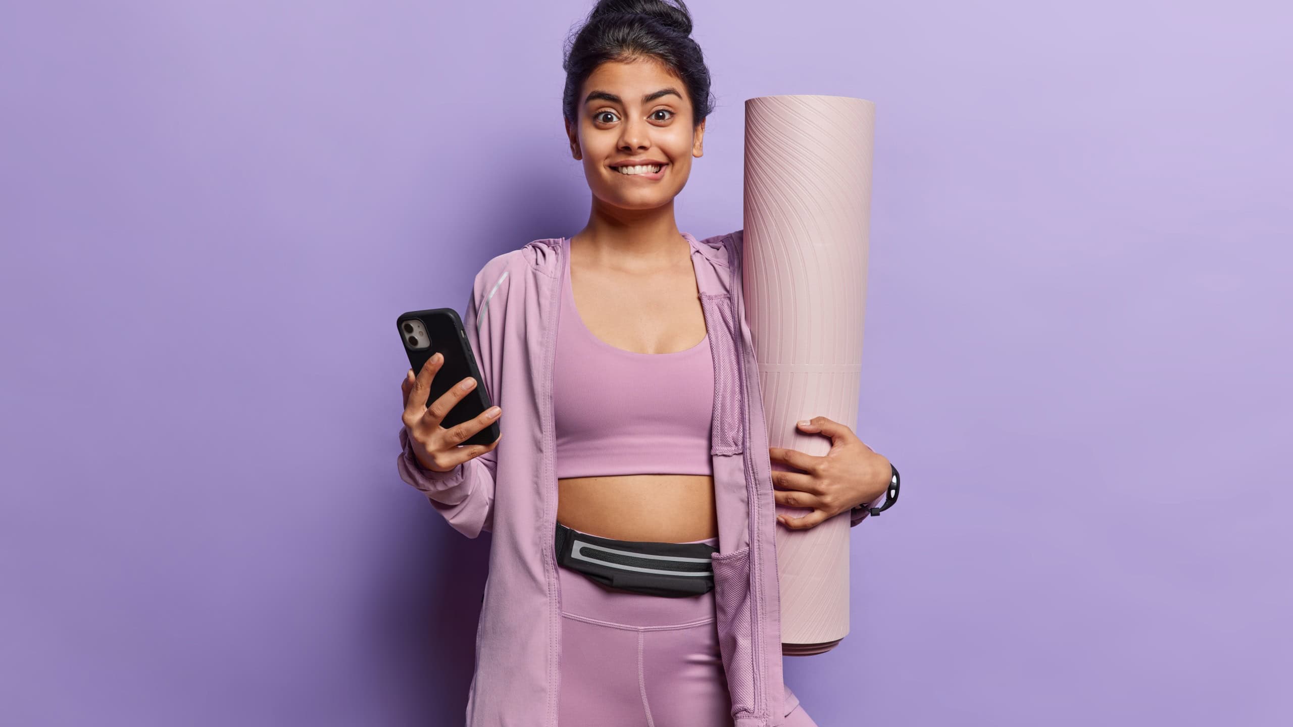 Indoor shot of beautiful dark haired sportswoman wearing sportswear poses with fitness mat in hand holds mobile phone finishes workout goes in for sport regularly isolated over purple background