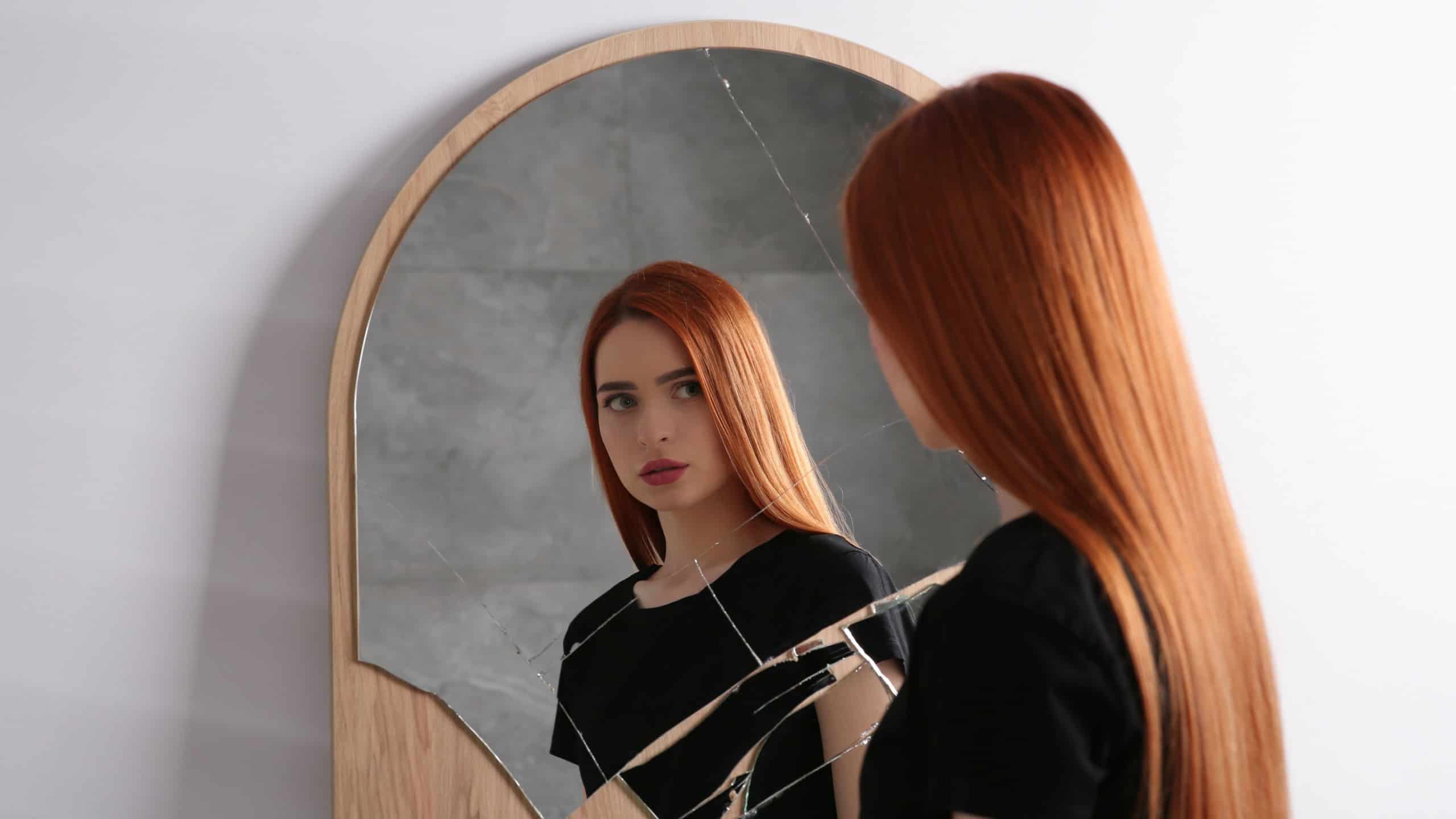 Mental problems. Depressed young woman looking at herself in broken mirror indoors