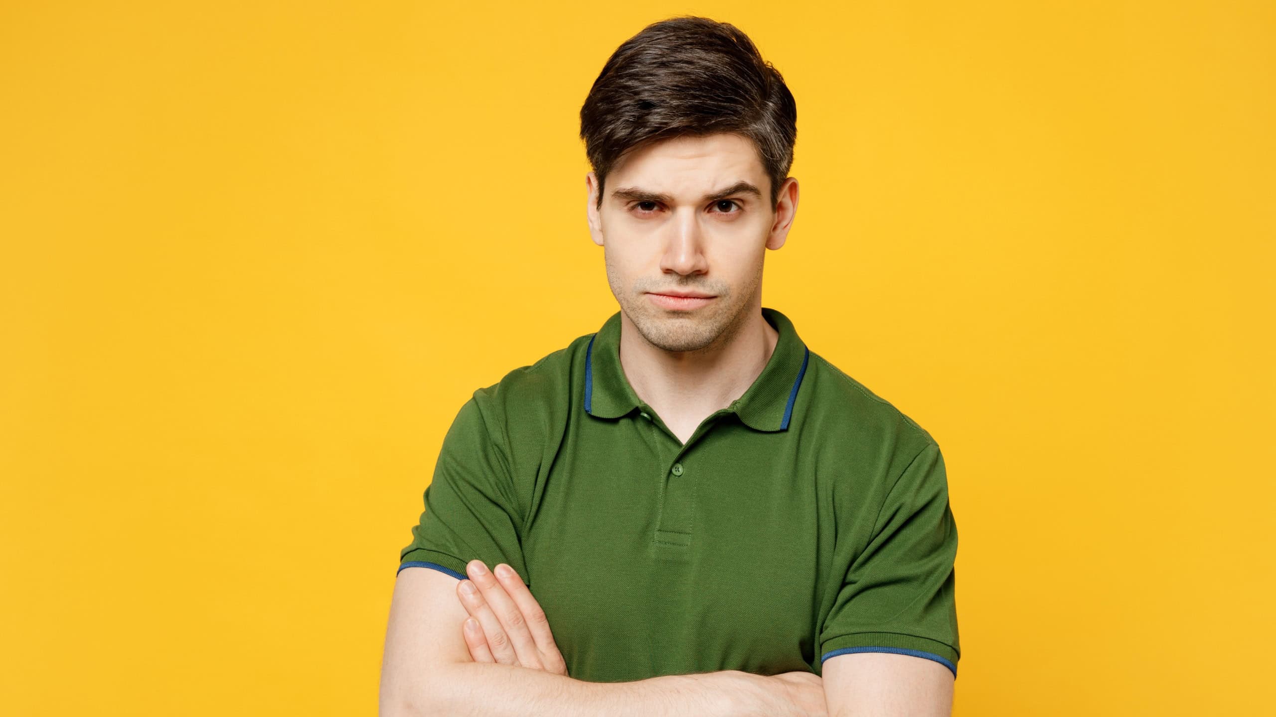 Young sad frowning grumoy serious caucasian man he wearing green t-shirt casual clothes holding hands crossed folded look camera isolated on plain yellow background studio portrait. Lifestyle concept