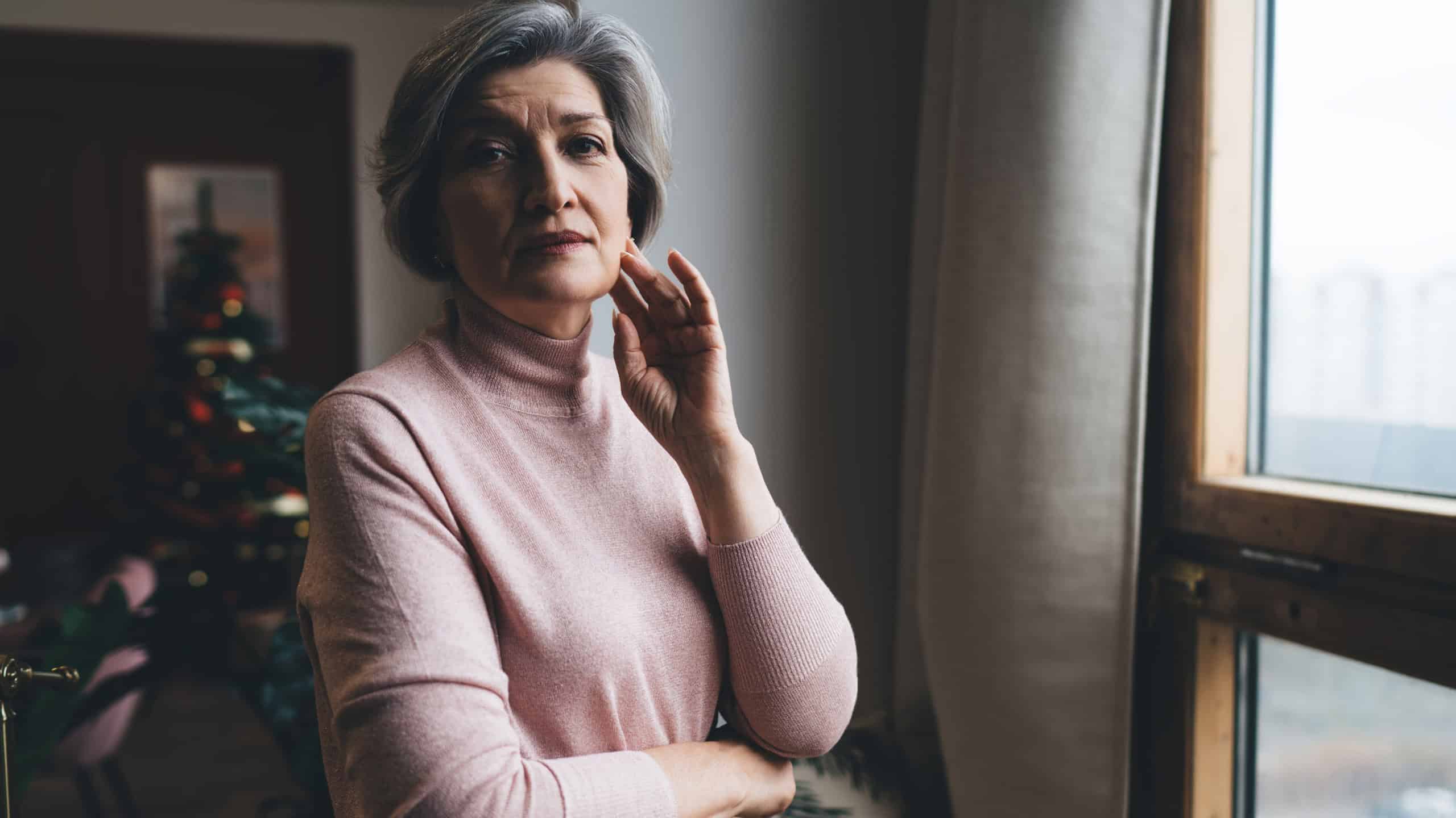 Lonely middle aged female with hand touching cheek looking at camera while standing near window with curtain in room with blurred Christmas tree with glowing lights at home