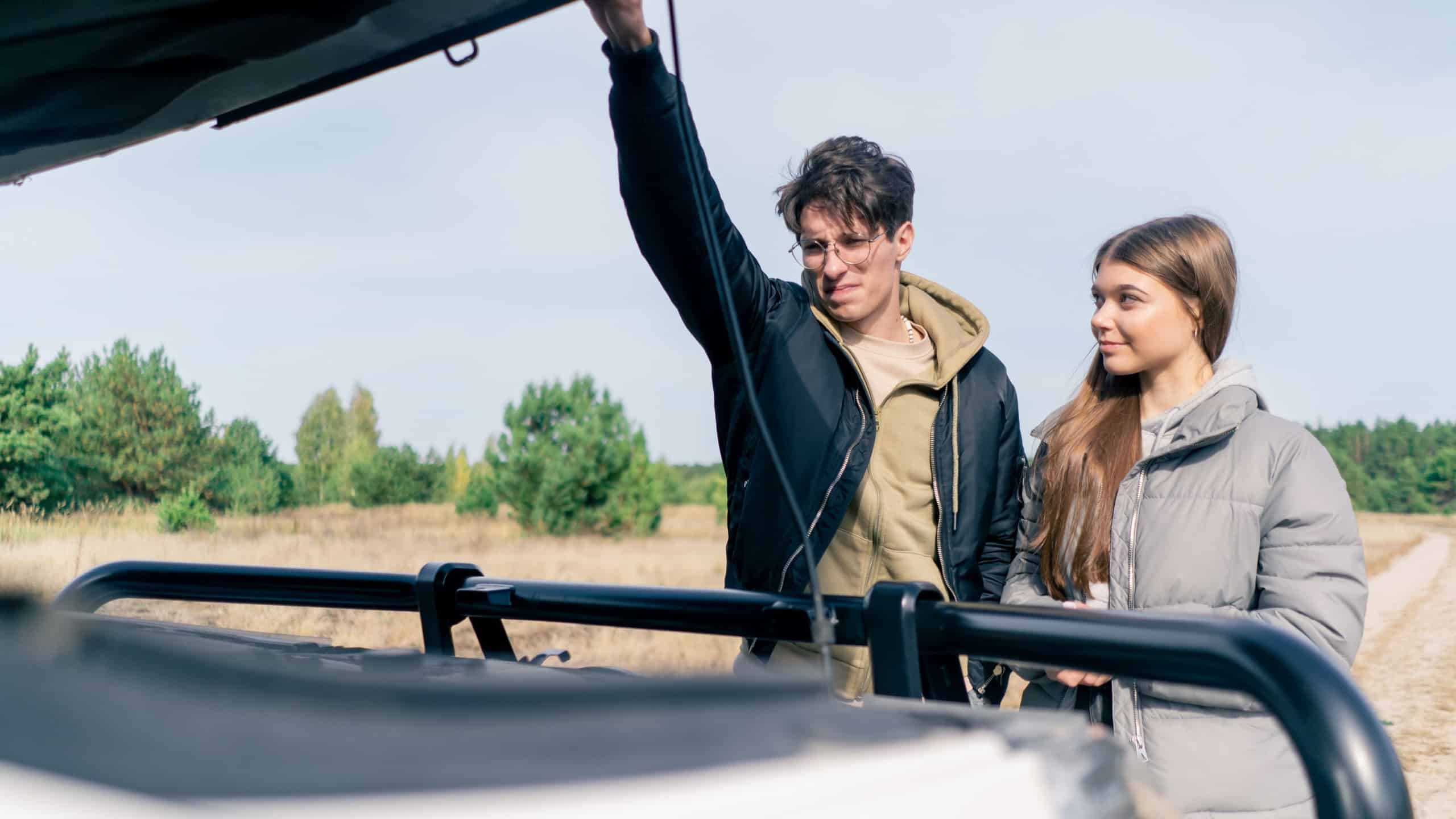 A guy driver helps a girl fix her car in a field and forest during a trip by opening the hood