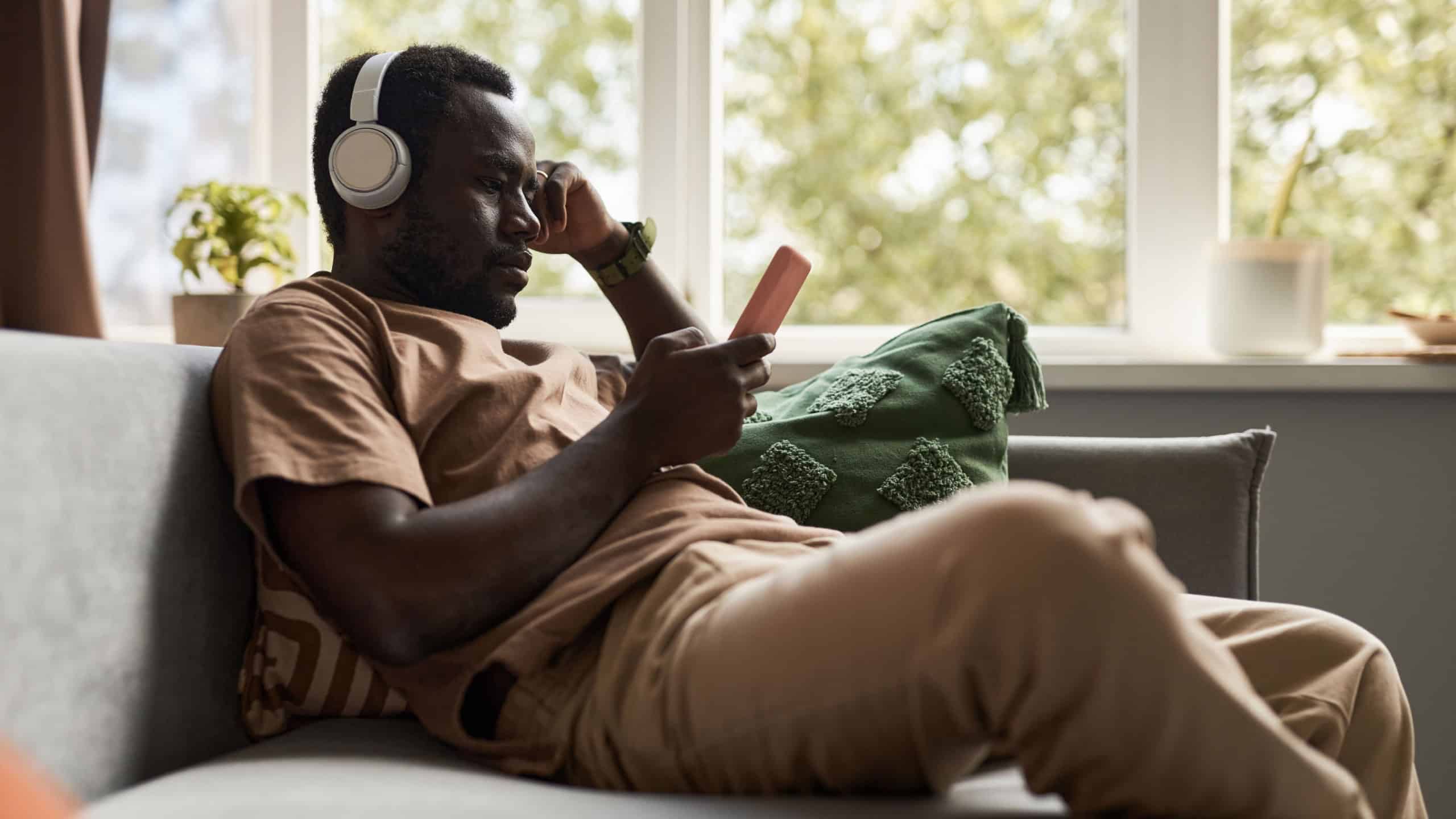 Side view portrait of African American man using smartphone with wireless headphones while listening to music on sofa at home