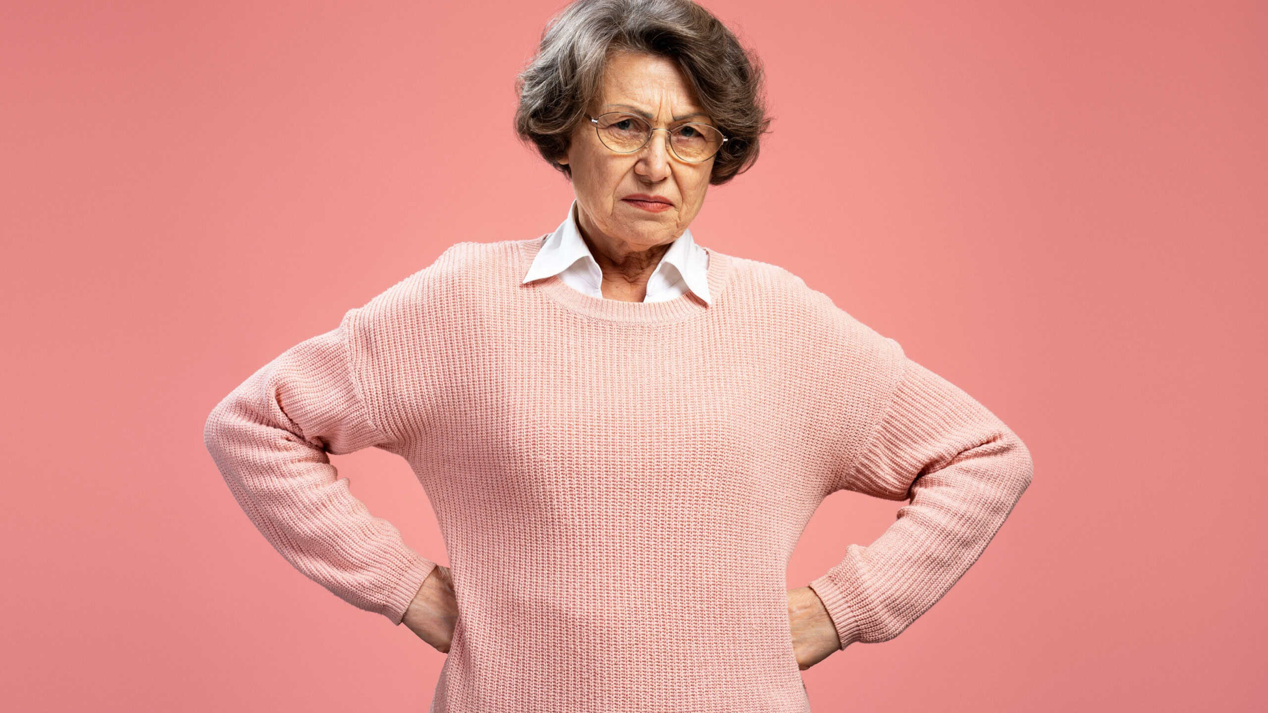 Angry serious gray haired 70 s woman wearing eyeglasses, hands on sides, looking at camera standing isolated on pink background. Bad mood concept
