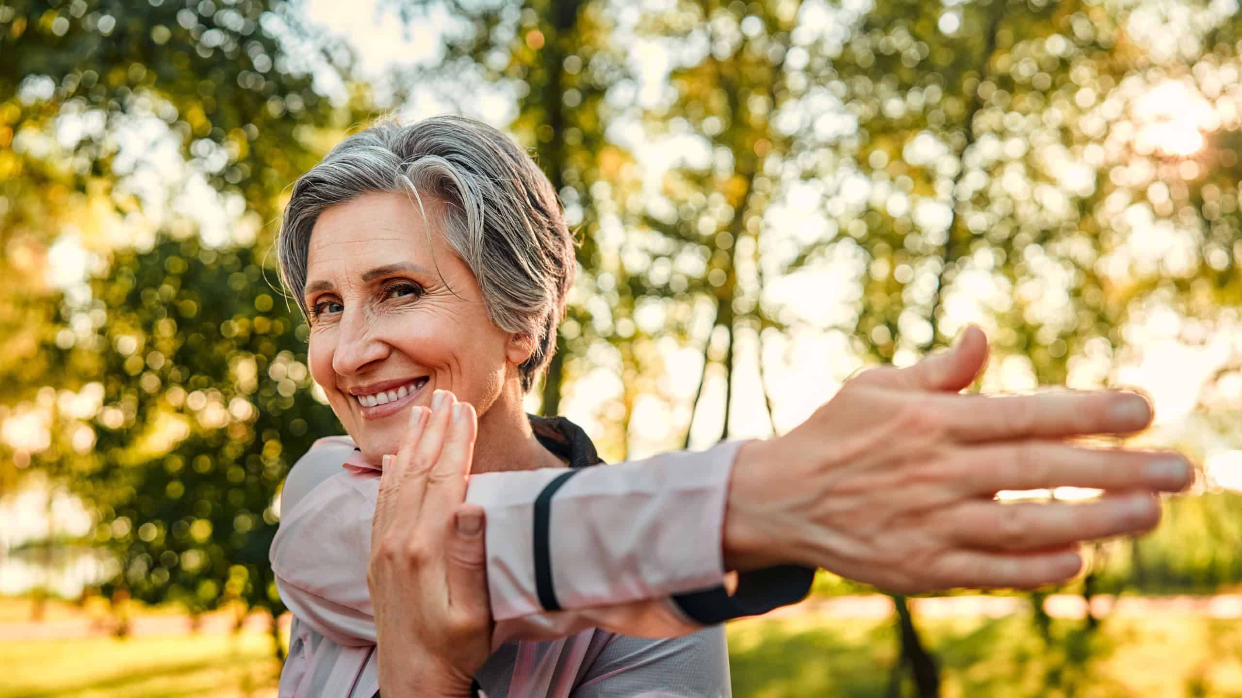 Active workout outdoors. Close up of pleasant grey haired woman in sportswear doing stretching exercises for arms at green sunny park. Caucasian lady of old ages taking care of health and body.
