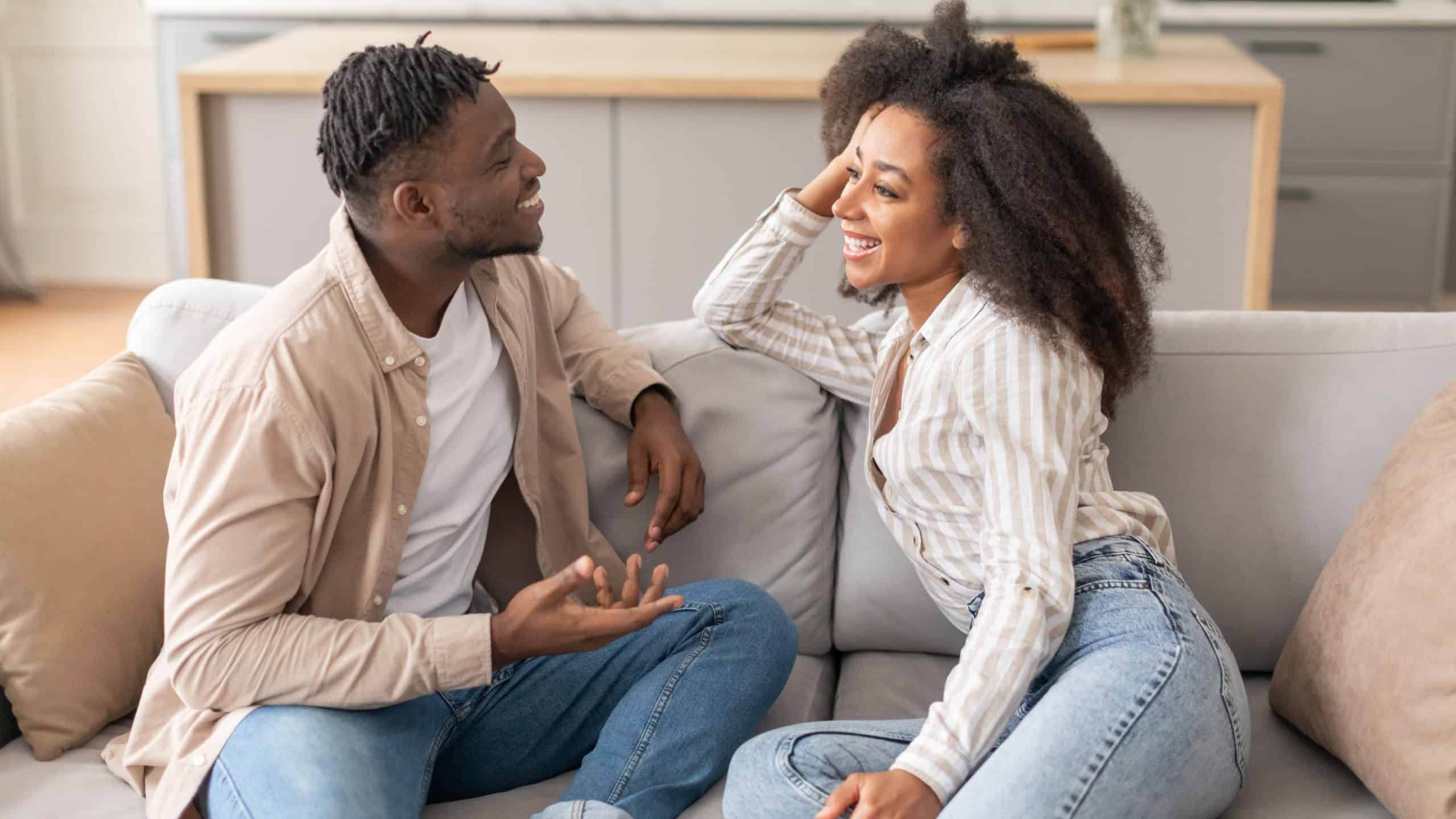 Happy Married African American Couple Talking Sitting On Sofa At Home On Weekend. Millennial Black Spouses Smiling To Each Other Enjoying Flirt And Conversation In Cozy Living Room