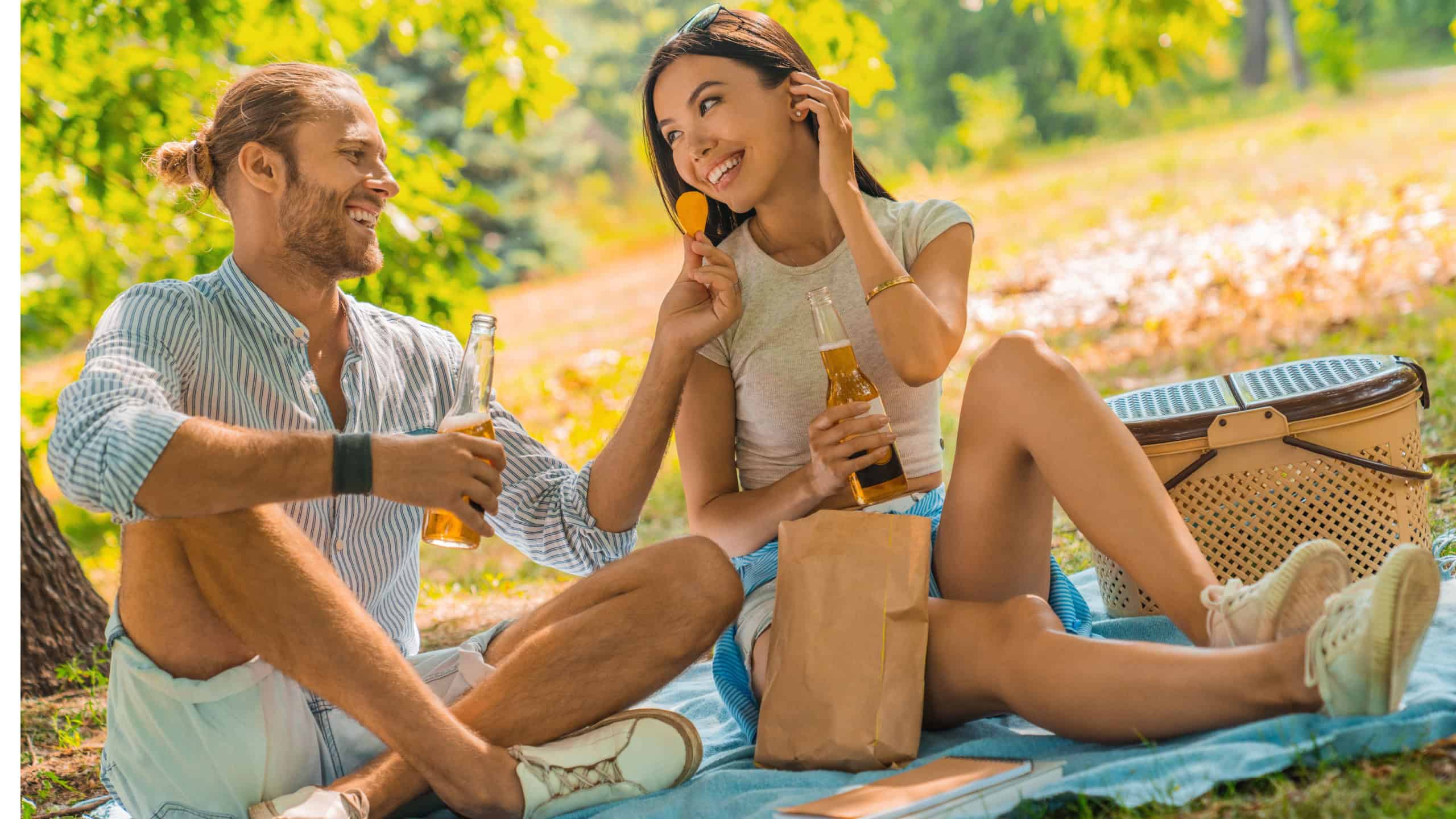 Young man offers a bit of chips to his girlfriend and they drinking beer. Couple enjoying their time together and are having a picnic at park on a romantic date