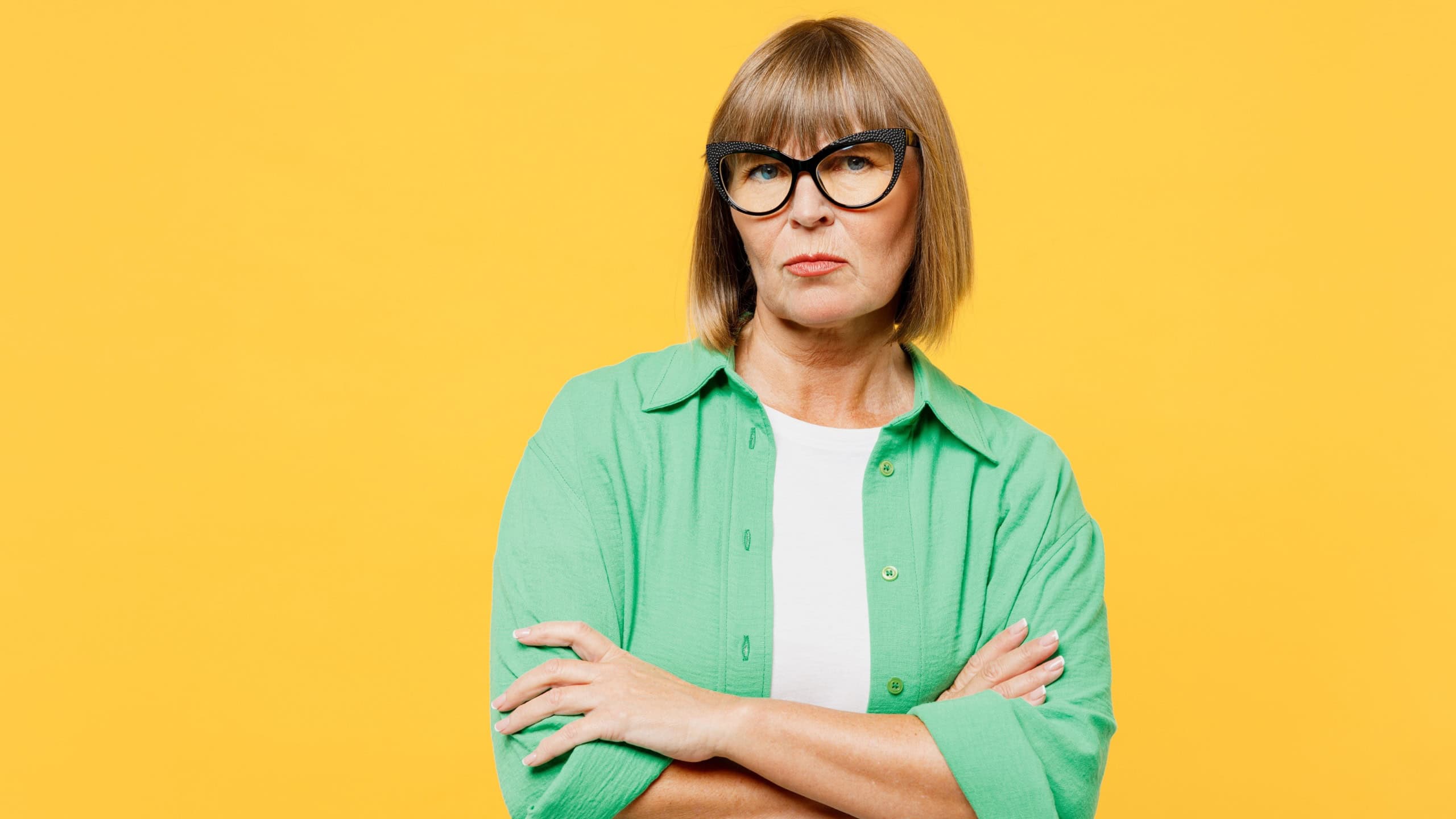 Elderly sad frowning suspicious blonde woman 50s years old wears green shirt glasses casual clothes hold hands crossed folded look camera isolated on plain yellow background studio. Lifestyle concept