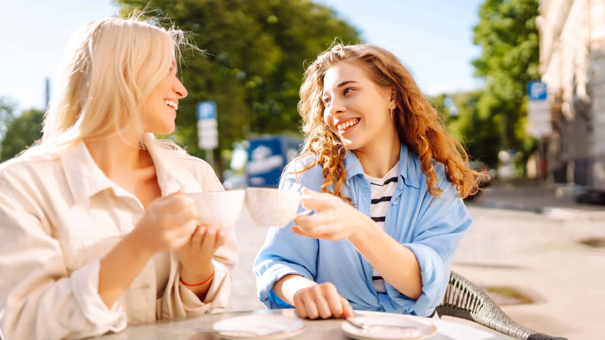 Portrait of a smiling young women friends sitting outdoors in cafe drinking coffee. Fashion, beauty, relax, tourism.