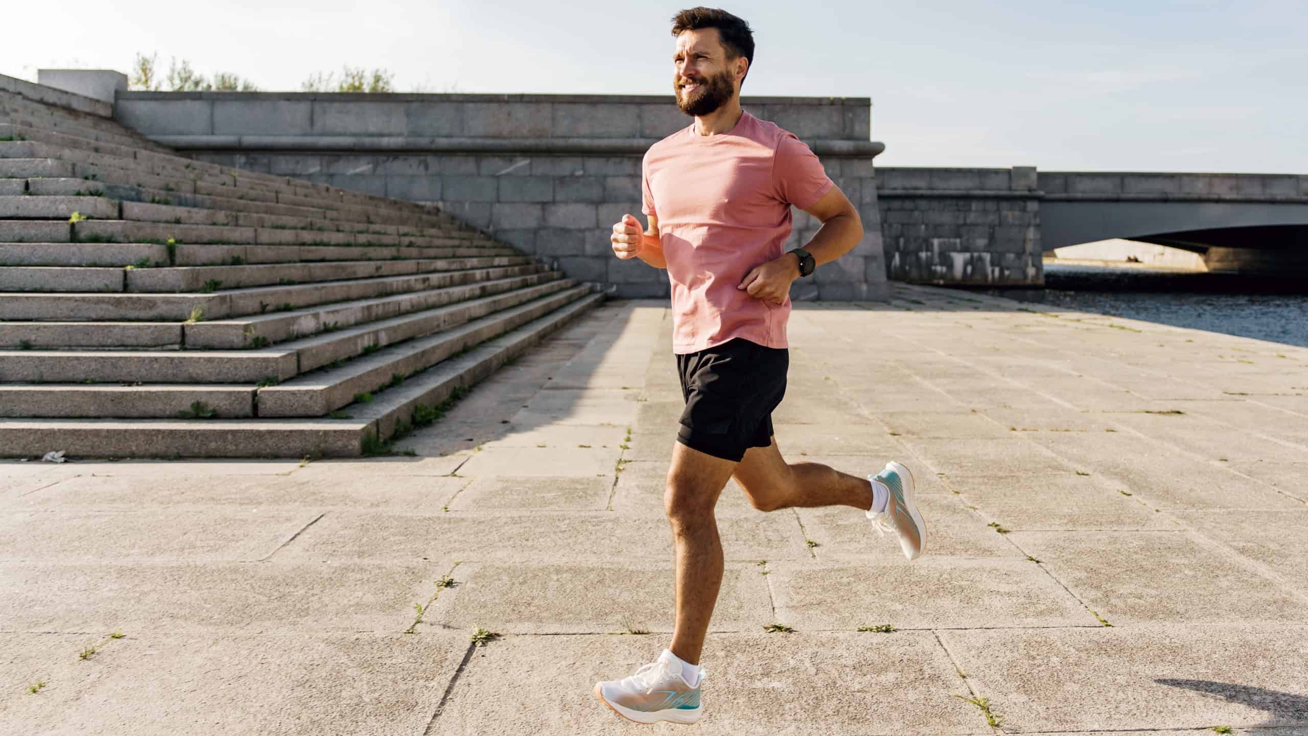 A man in a pink shirt and black shorts runs with purpose along the riverside promenade, with wide steps and a bridge in the background.