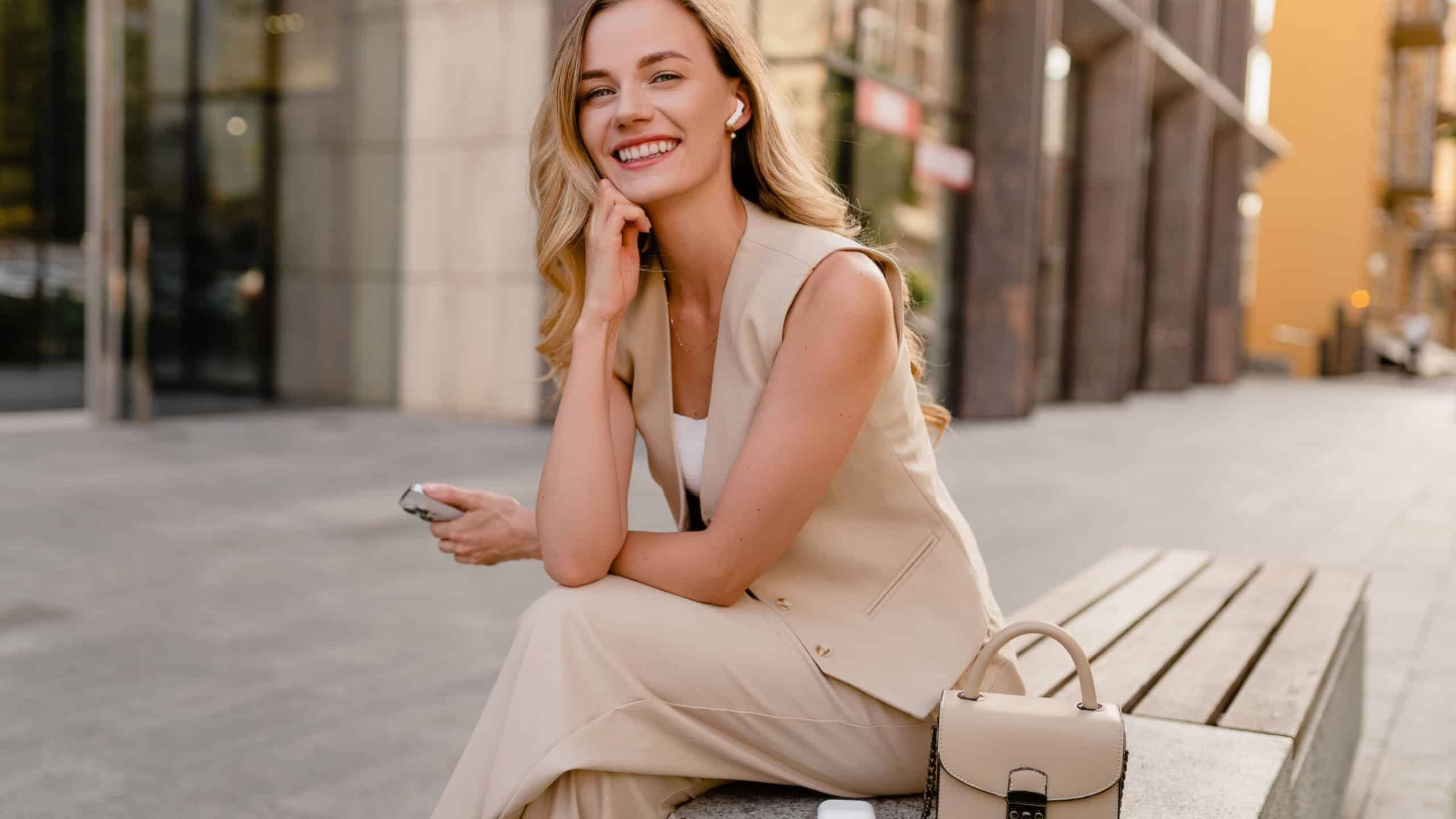 attractive young blond woman sitting in city street using smart phone in spring summer autumn fashion trend outfit smiling happy wearing beige suit and handbag style in sunny warm tones
