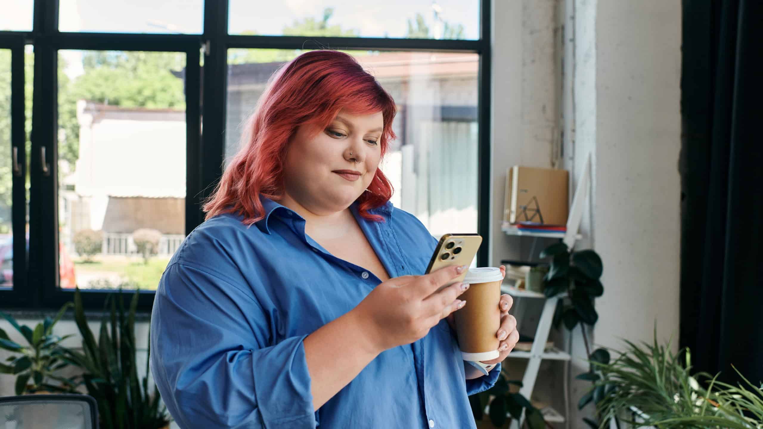 A plus size businesswoman with vibrant pink hair checks her phone while holding a cup of coffee.
