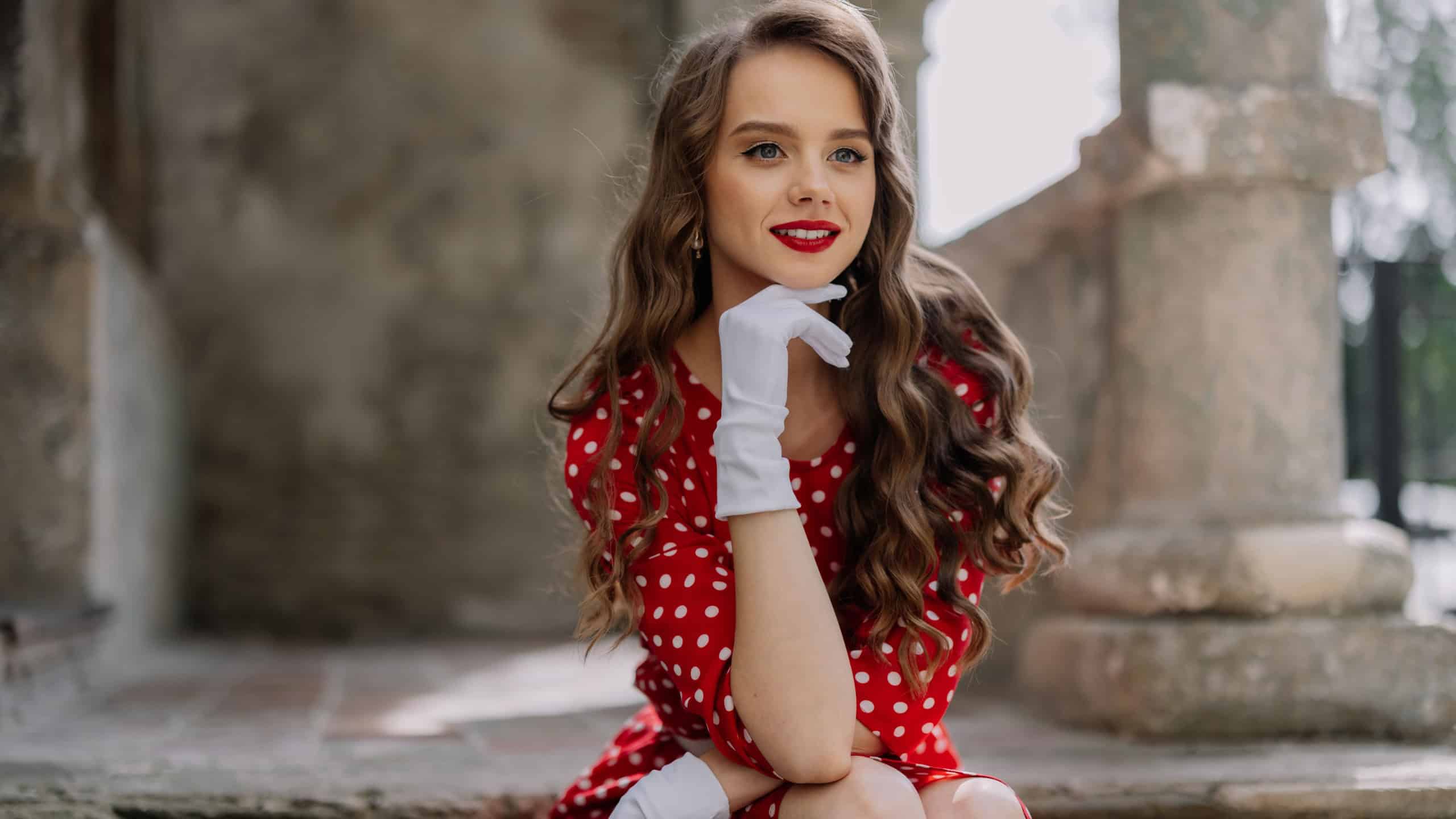 Stylish woman wearing red polka dot dress and white gloves sits gracefully on stone steps, evoking vintage charm and elegance