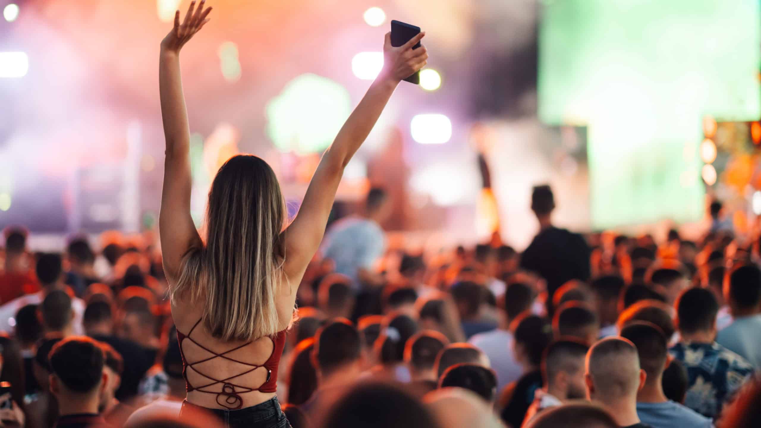 A woman in a vibrant festival crowd raises her phone to capture the ecstatic atmosphere, surrounded by lights, music, and people enjoying the event.