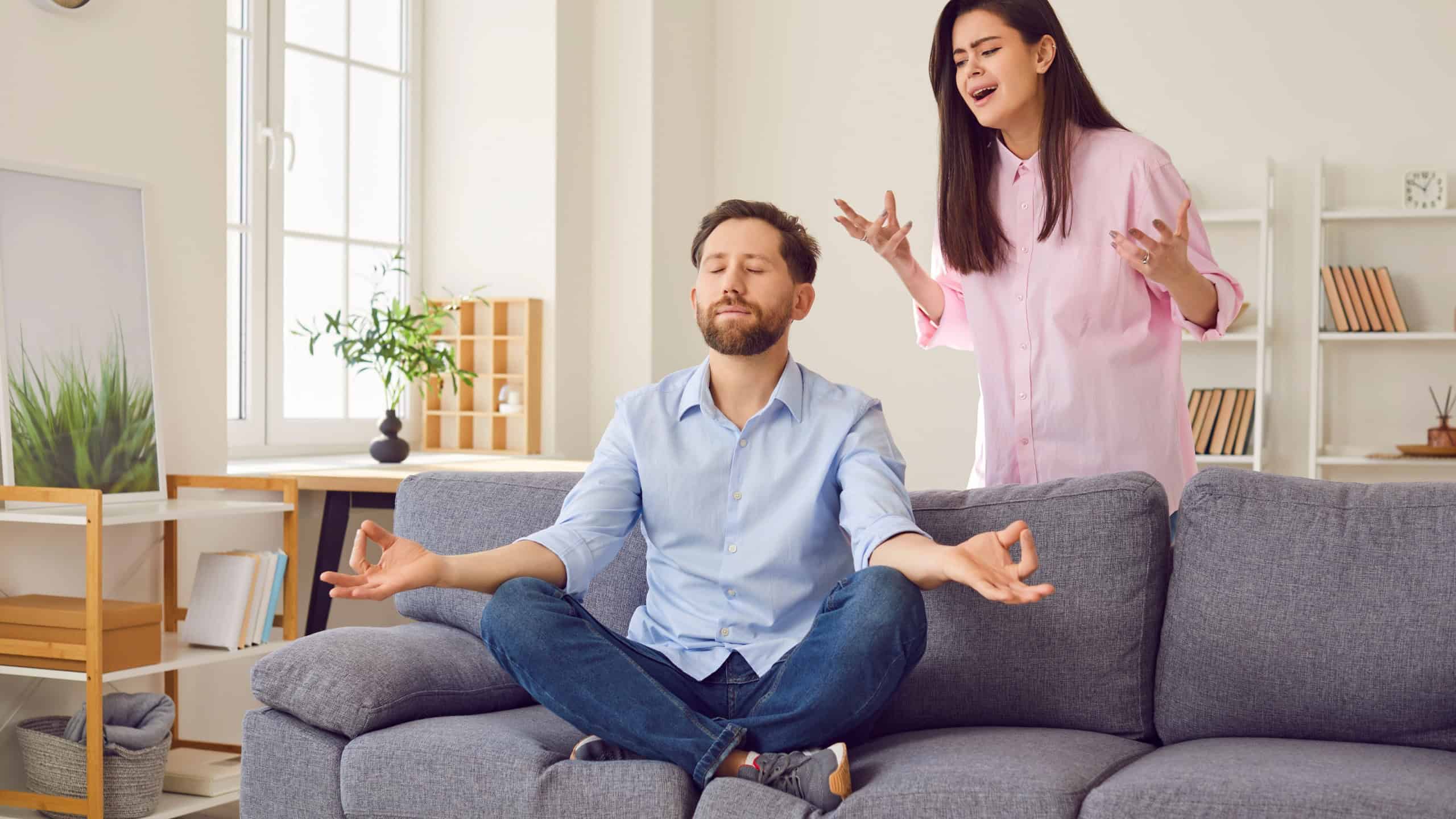 Upset and annoyed woman yelling at man who is meditating and ignoring problem. Man with eyes closed is sitting in zen pose, while woman is standing next to him with negative expression on her face.