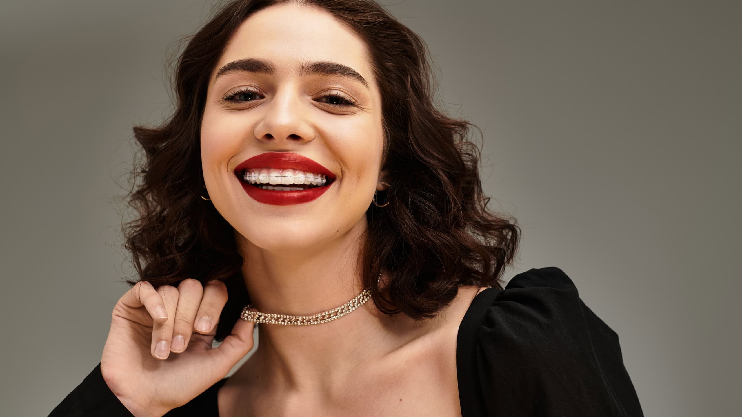 A confident young woman shows off her braces and vibrant red lips against a grey backdrop.